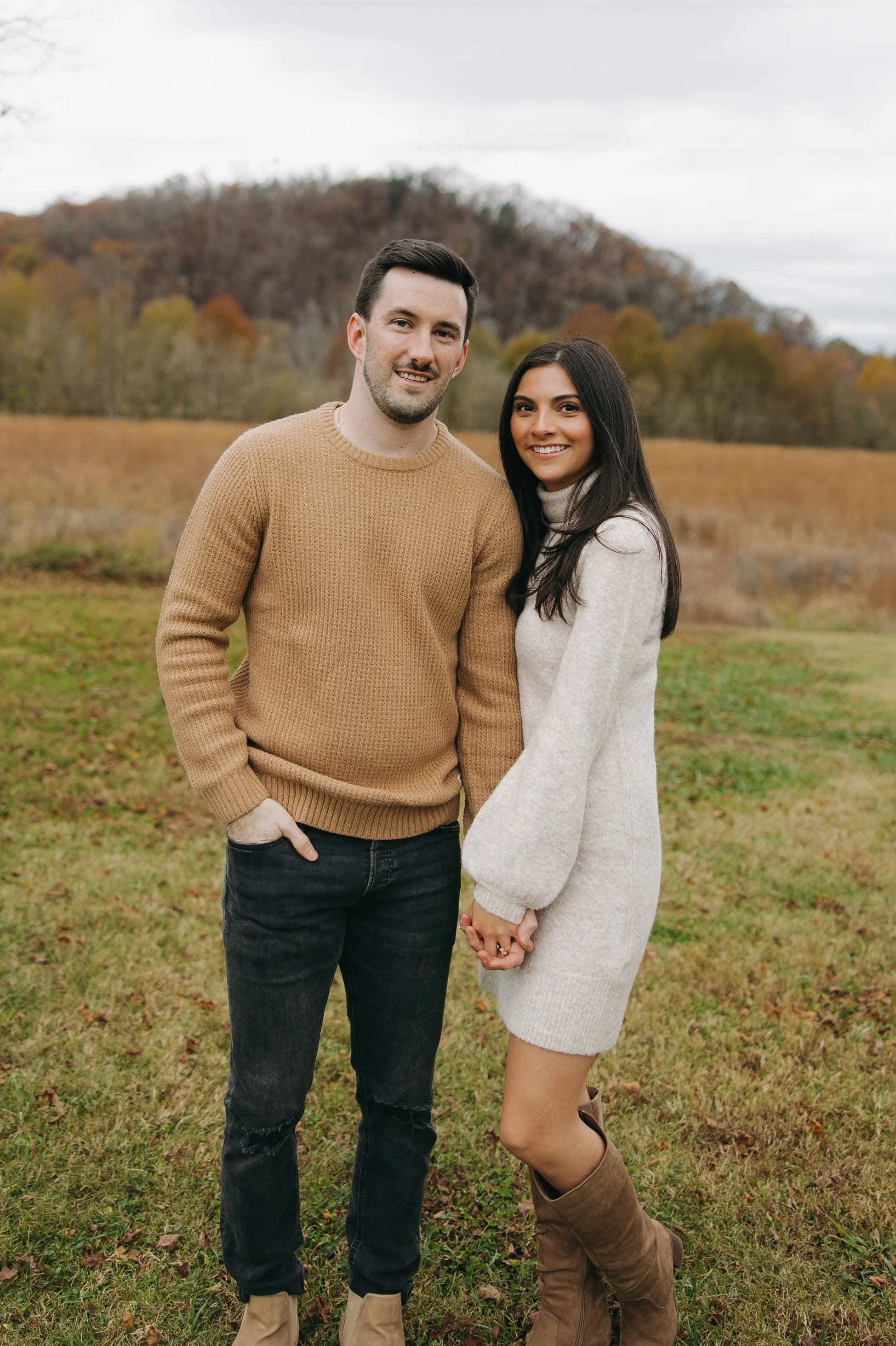 A young couple holding hands outdoors in an autumn landscape with trees and a hill in the background. The man has dark hair, a beard, and is wearing a beige sweater, black jeans, and tan boots. The woman has long dark hair and is wearing a light-colored sweater dress and brown boots.