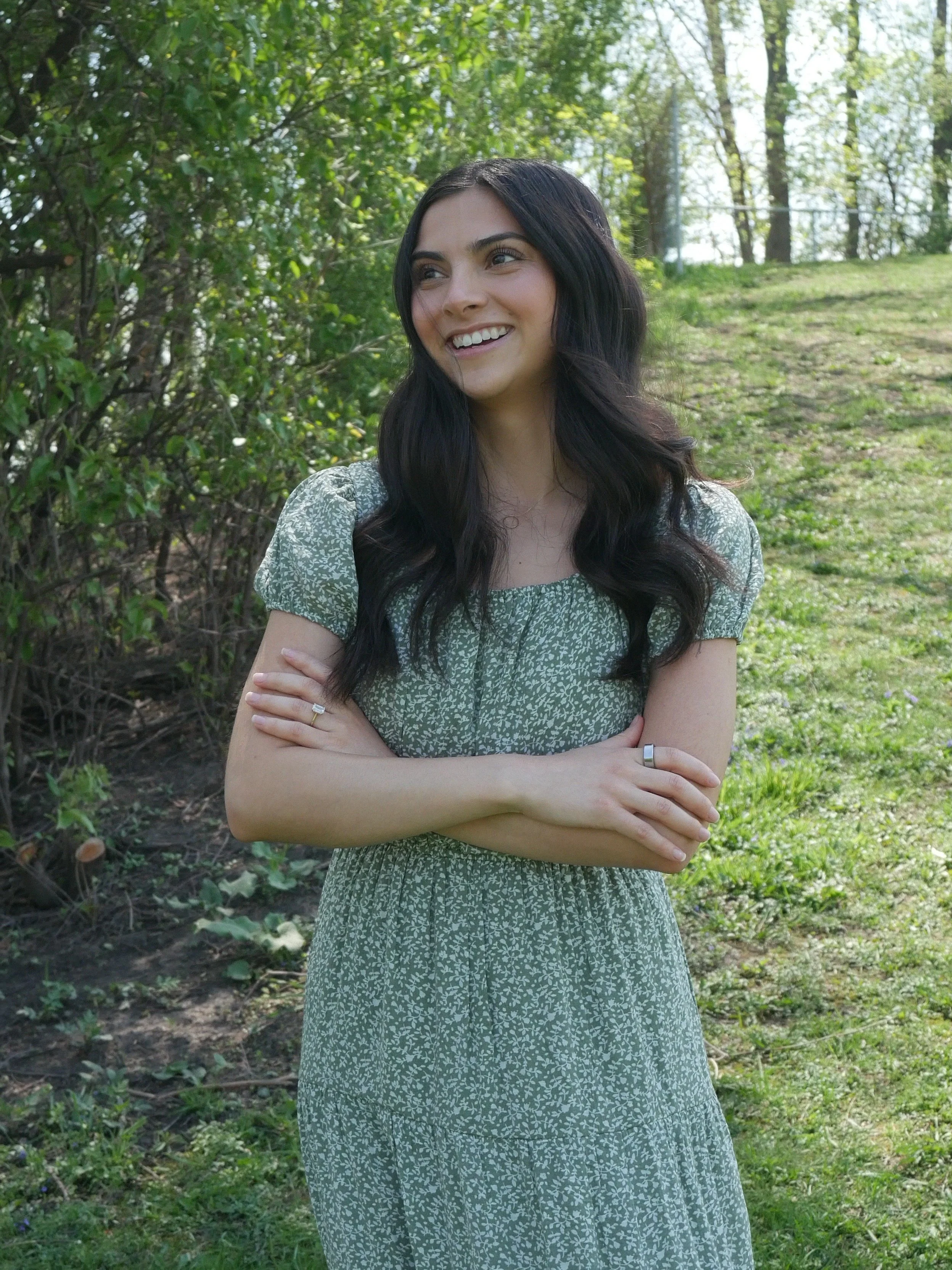 A woman with long, dark hair wearing a green, short-sleeved, floral dress stands outdoors in a grassy area with trees in the background. She is smiling and has her arms crossed.
