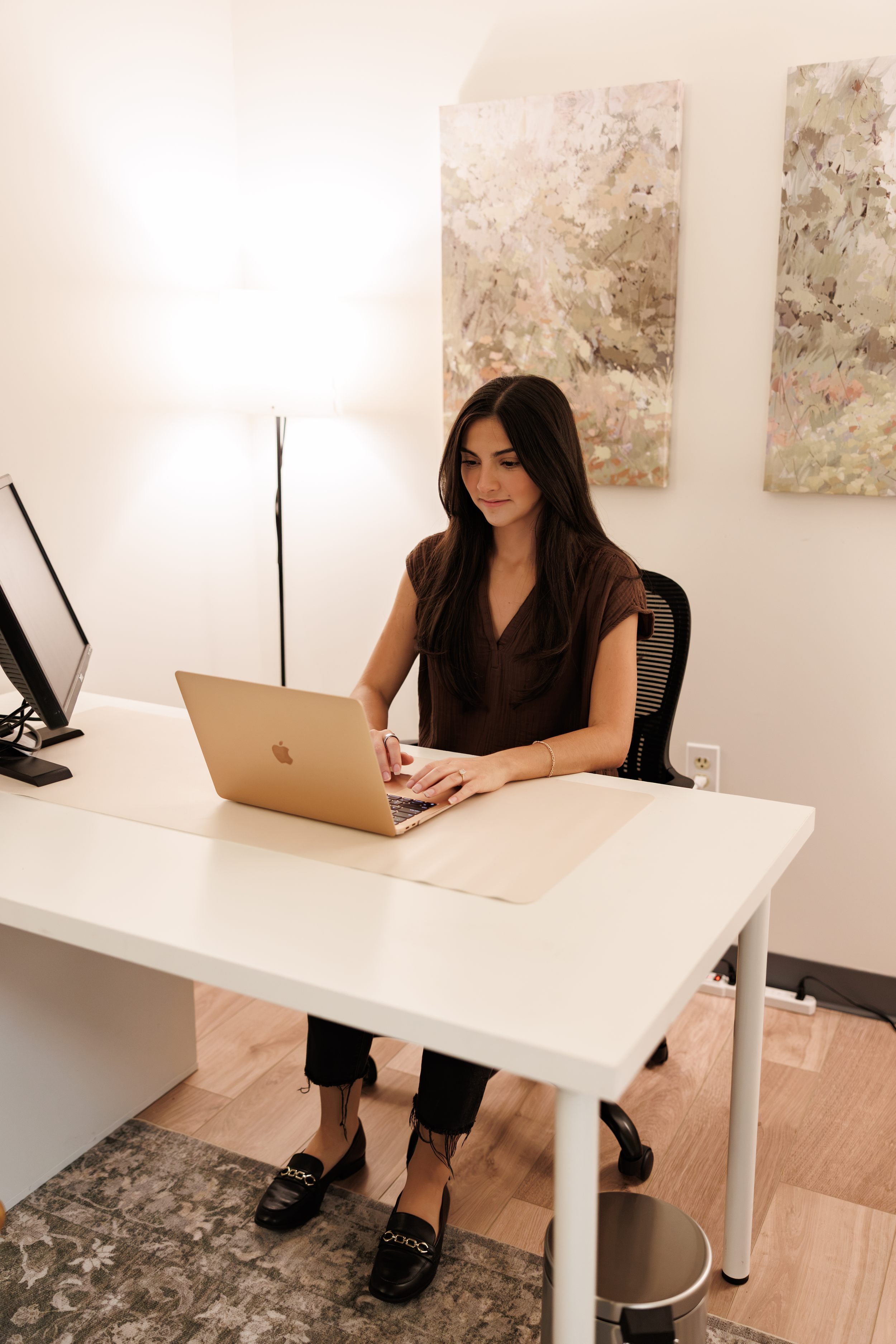 A woman with long dark hair sitting at a white desk working on a silver laptop in a modern office. There is a computer monitor on her left, a standing lamp providing light, and abstract artwork hanging on a white wall behind her. She is wearing a brown top, black ripped jeans, and black loafers with gold chain details.