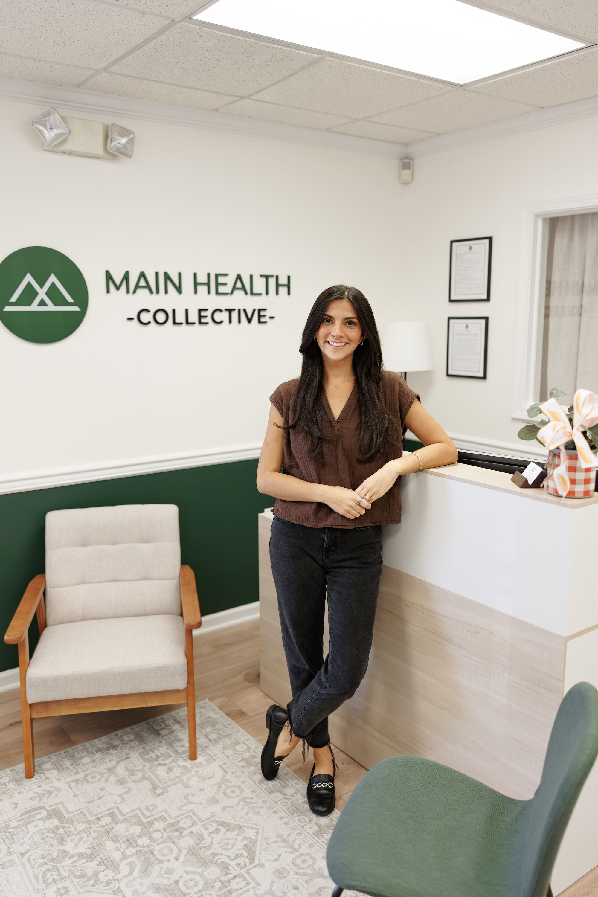 A woman with long dark hair, wearing a brown top and black pants, standing at a reception desk in a medical office. Behind her, a wall features the logo and name "Main Health Collective." There are framed documents on the wall, a white lamp, and a desk with flowers, and the office has a beige chair and green upholstered chairs, with a patterned carpet on the floor.