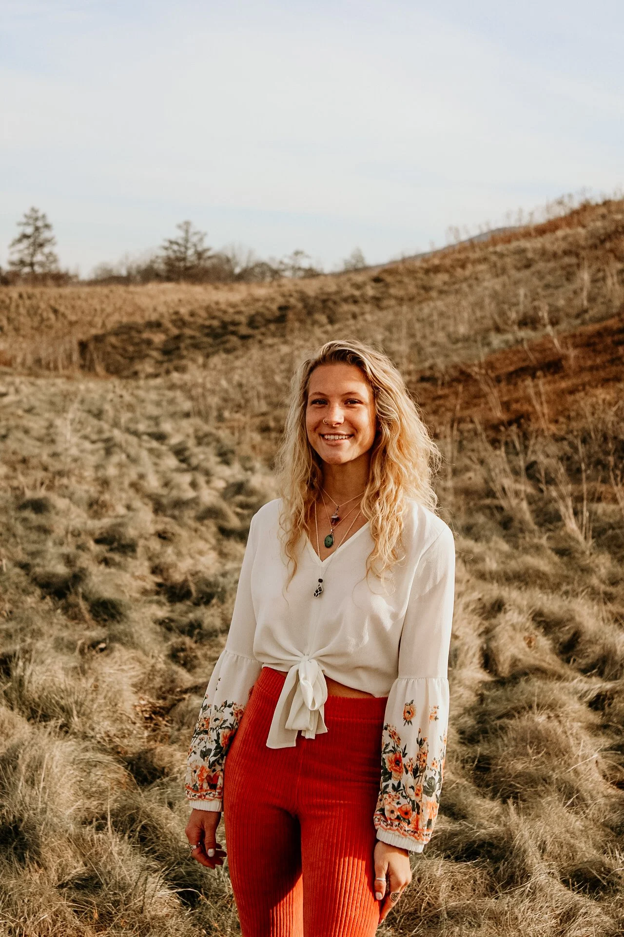 A young woman with blonde curly hair and wearing a white blouse with embroidered sleeves and orange pants, smiling outdoors in a countryside setting with dry grass, hills, and sparse trees in the background.