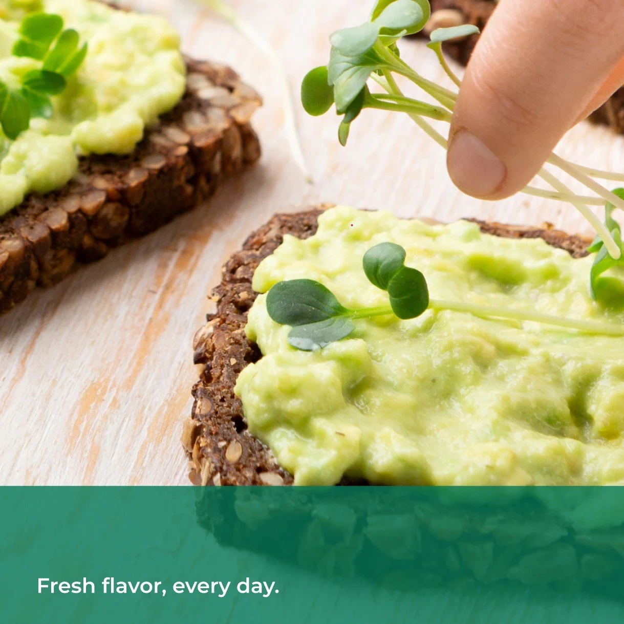 Close-up of avocado toast on seeded bread topped with fresh microgreens, with a hand placing additional greens on top. Text overlay reads “Fresh flavor, every day.”