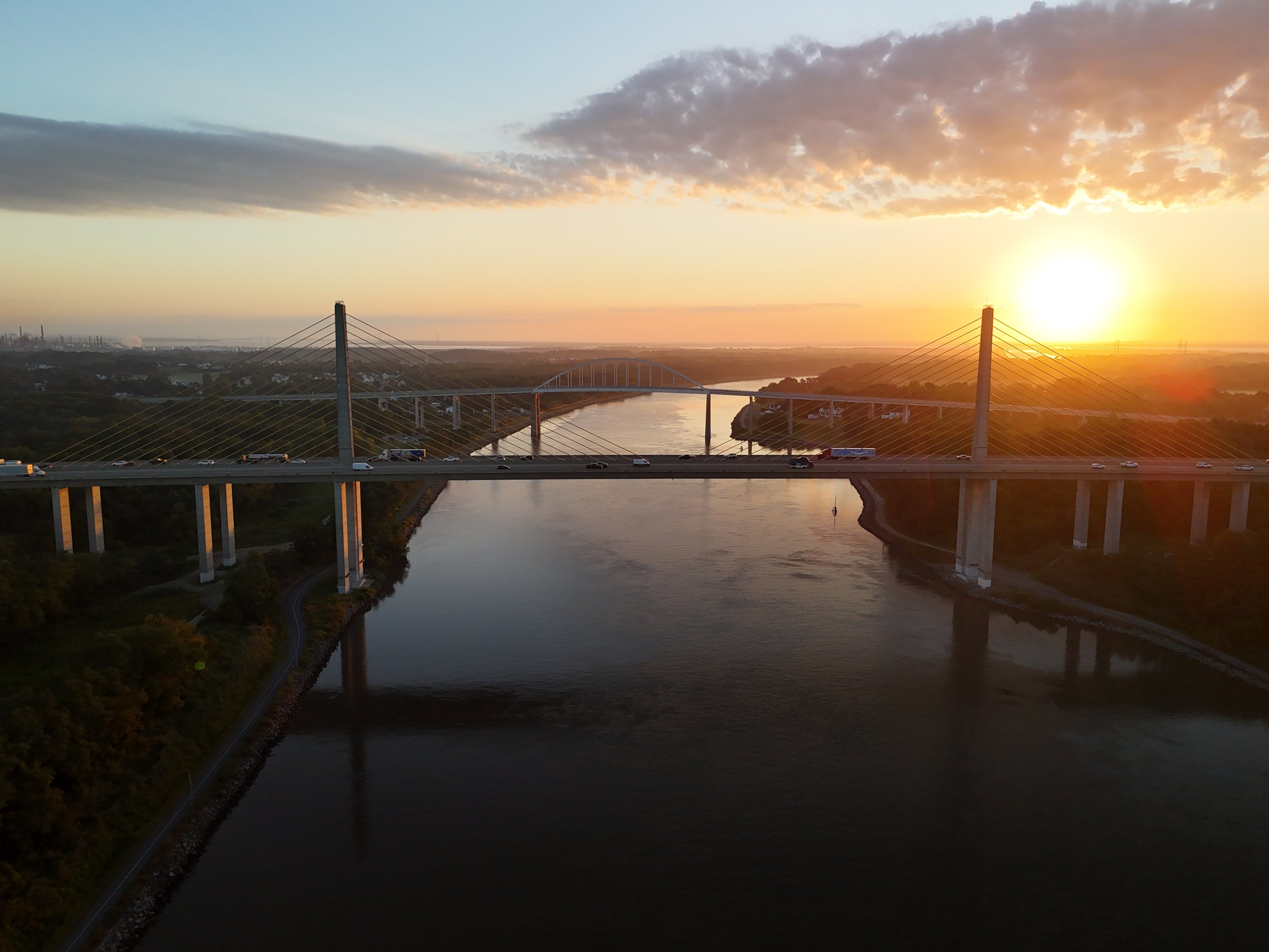 Aerial view of a large bridge with tall pylons and cables over a river at sunset, with cars traveling across, and the sun near the horizon illuminating the sky with orange and purple hues.