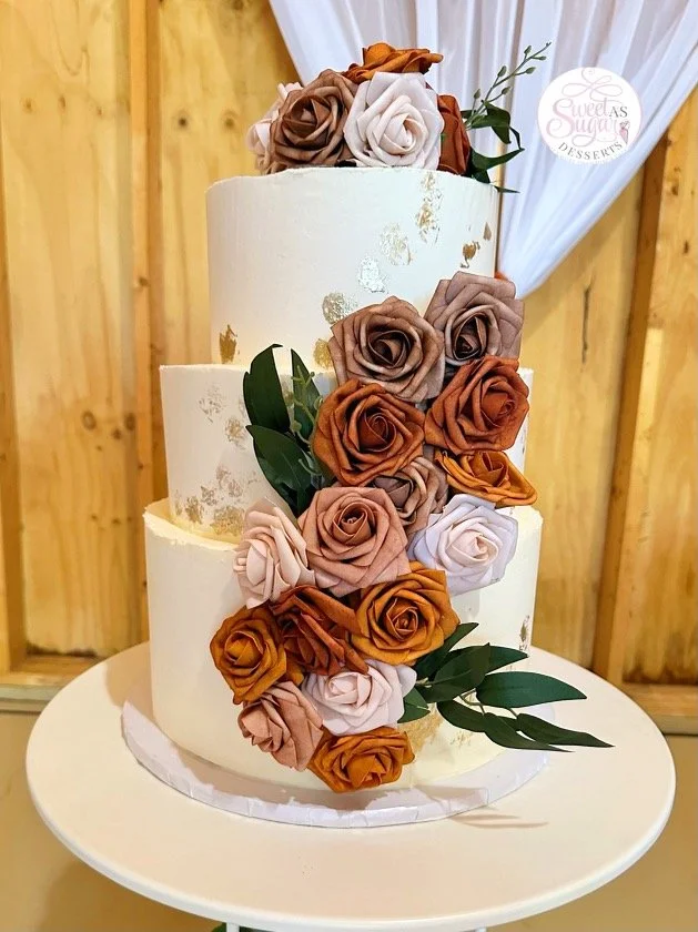 Three-tiered white wedding cake decorated with caramel, blush, and taupe roses and green leaves, with gold accents and a wooden background.