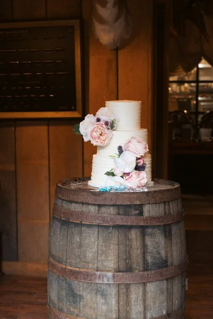 White tiered wedding cake with pink and white flowers on a wooden barrel in a rustic setting.