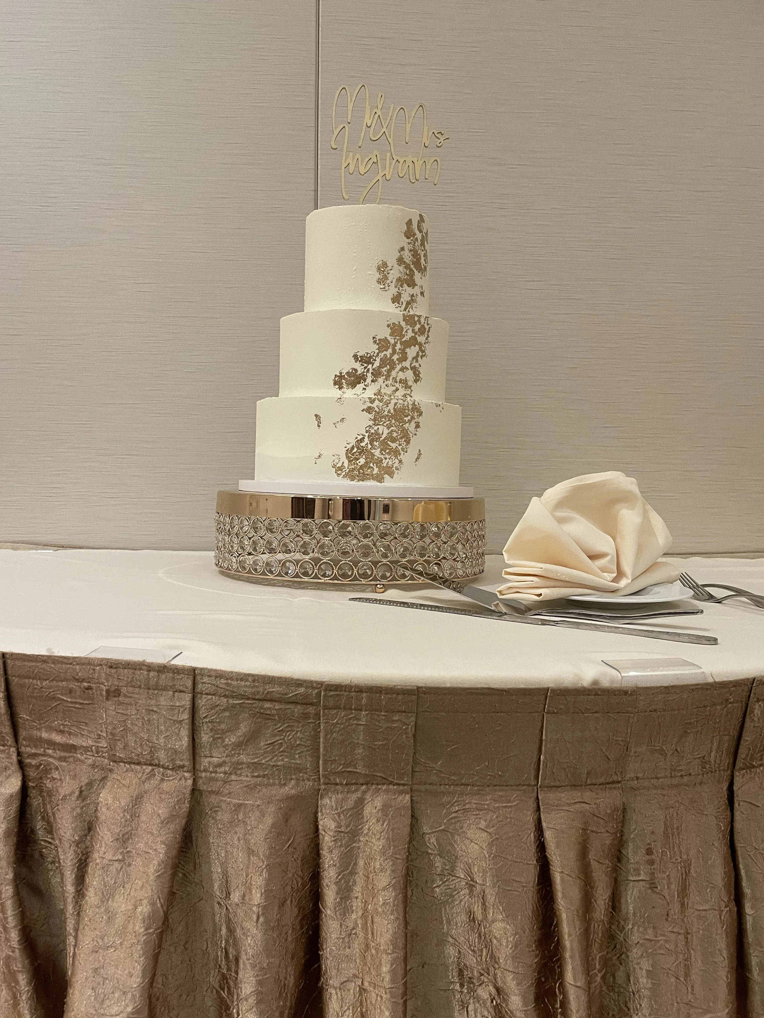 A three-tier wedding cake with gold leaf decoration, placed on a sparkling silver cake stand. There is a folded napkin, a fork, and a knife beside it, on a table draped with cream-colored cloth. A wedding topper reads 'Mr & Mrs Nguyen'.
