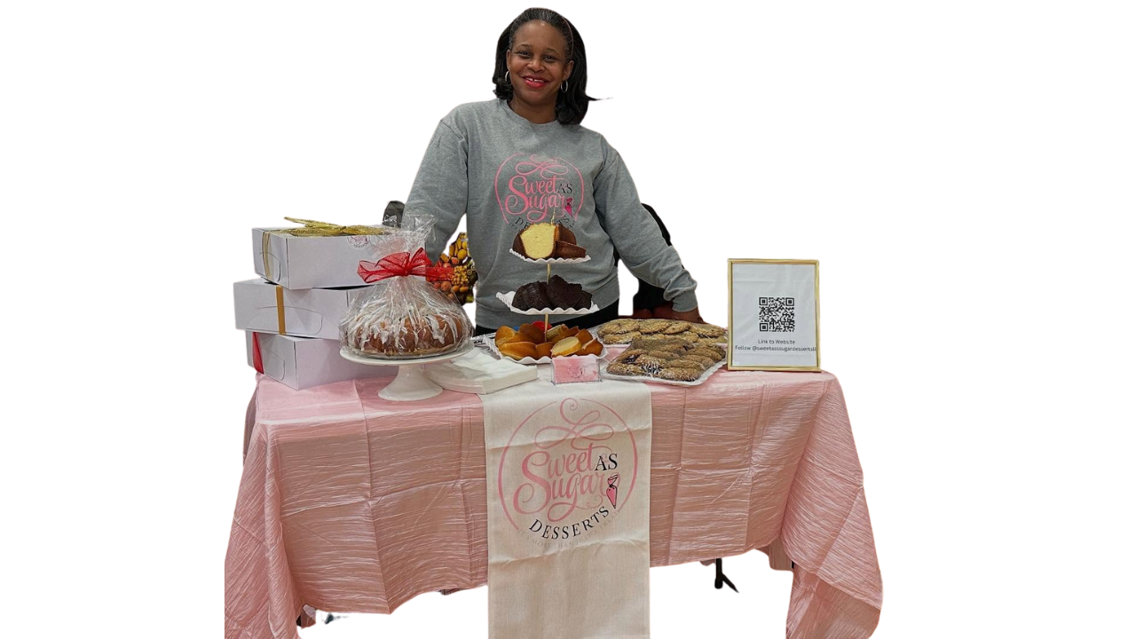 A woman standing behind a table with baked goods and desserts, wearing a gray sweatshirt with a pink logo, on a pink tablecloth with a "Sweet as Sugar" banner.