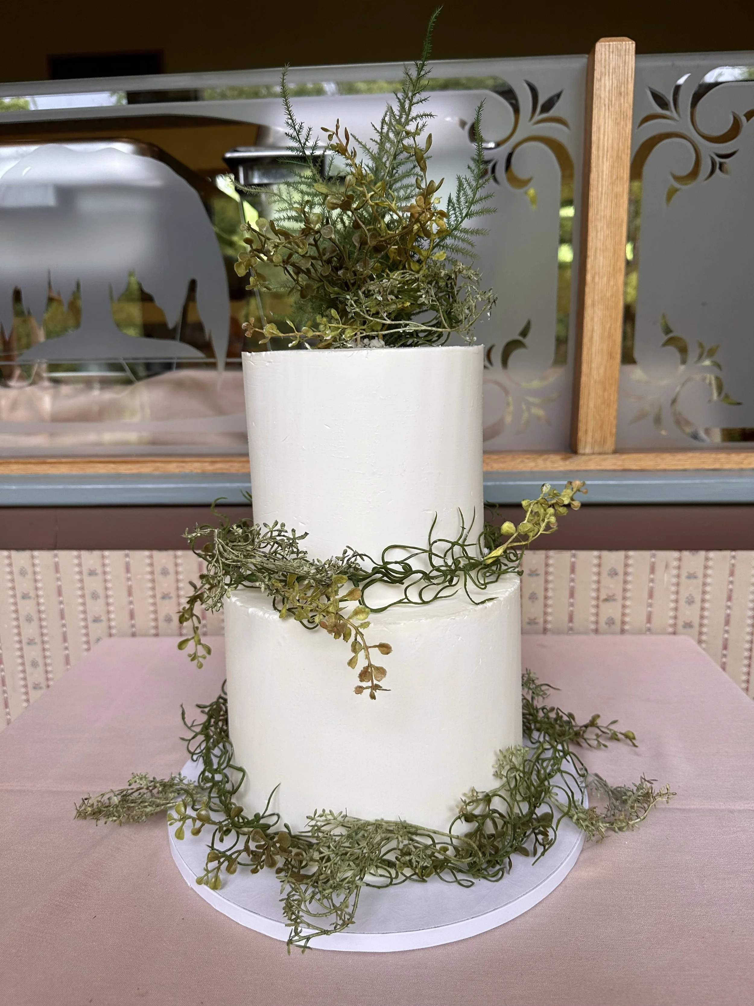Two-tier white wedding cake decorated with greenery and succulents, placed on a pink tablecloth.