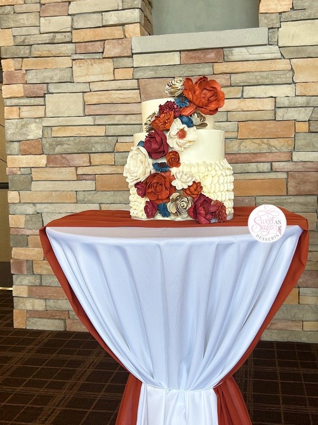 A tiered wedding cake decorated with colorful sugar flowers, placed on a table with a white and rust-colored draped cloth against a stone wall background.