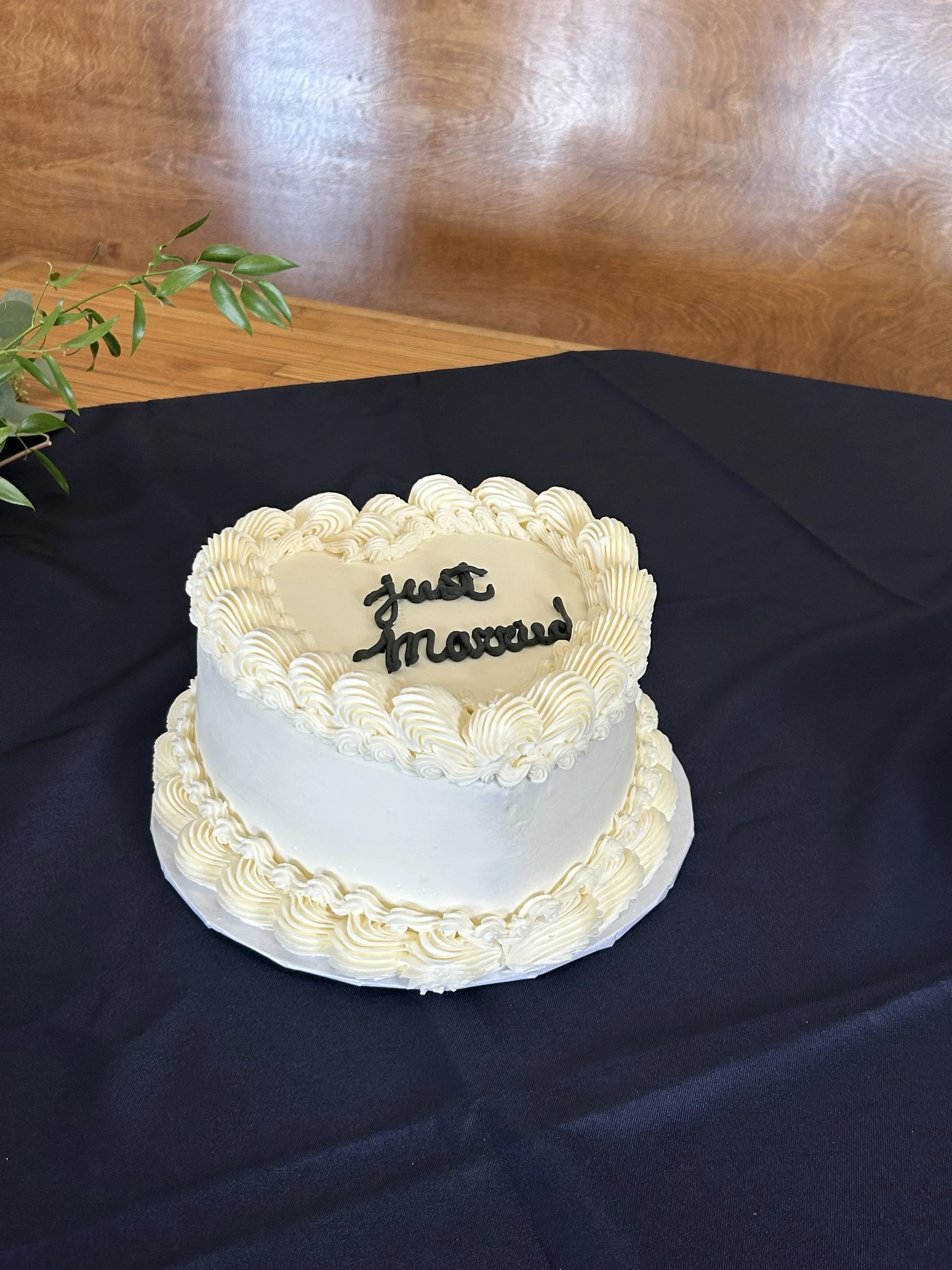A white wedding or celebration cake with piped cream decoration on top and around the base. The cake has a message that reads 'Just Married' in black icing on the top.