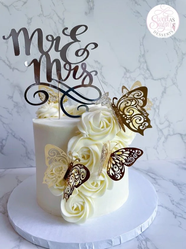 Wedding cake with white roses and gold and brown butterfly decorations, topped with a "Mrs & Mrs" cake topper, on a white cake board against a marble background.