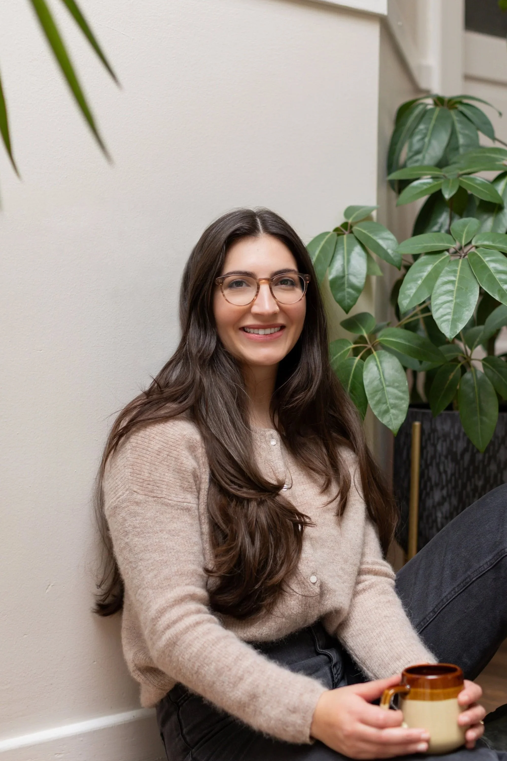 A woman with long brown hair and glasses sitting against a beige wall, holding a mug with a large green plant nearby.
