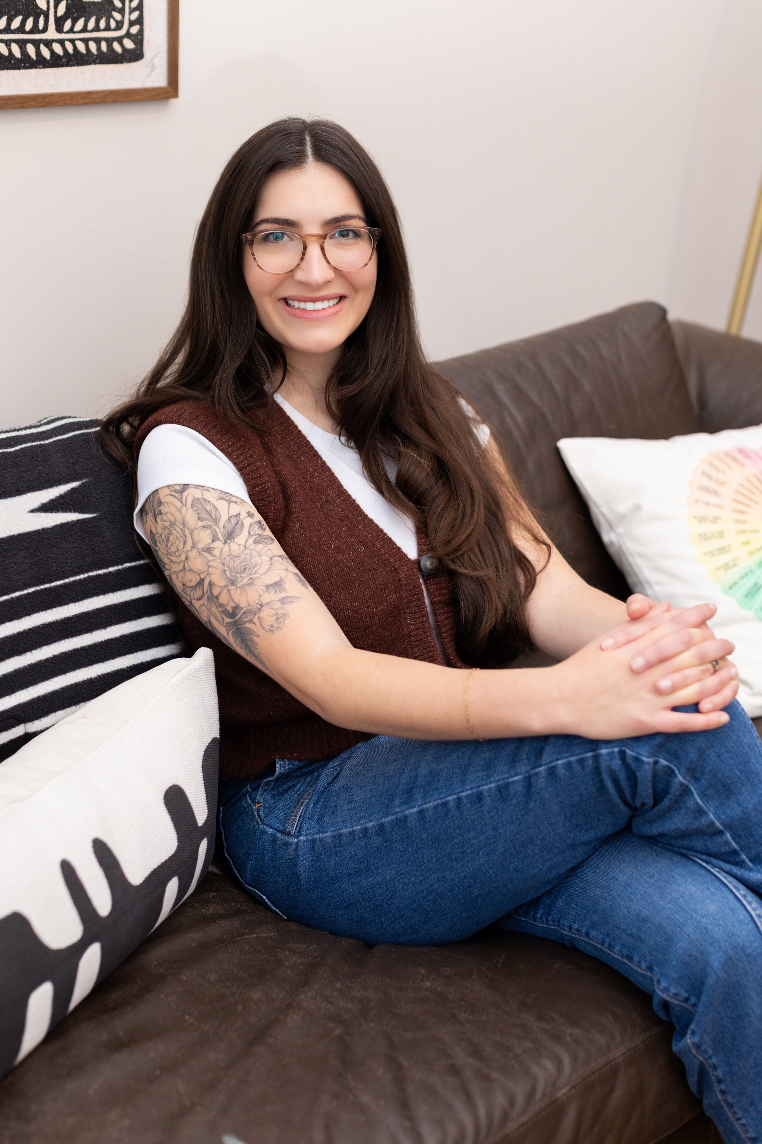 A young woman with long brown hair, glasses, and a tattoo on her left arm, smiling while sitting on a brown leather couch with decorative pillows in a living room.