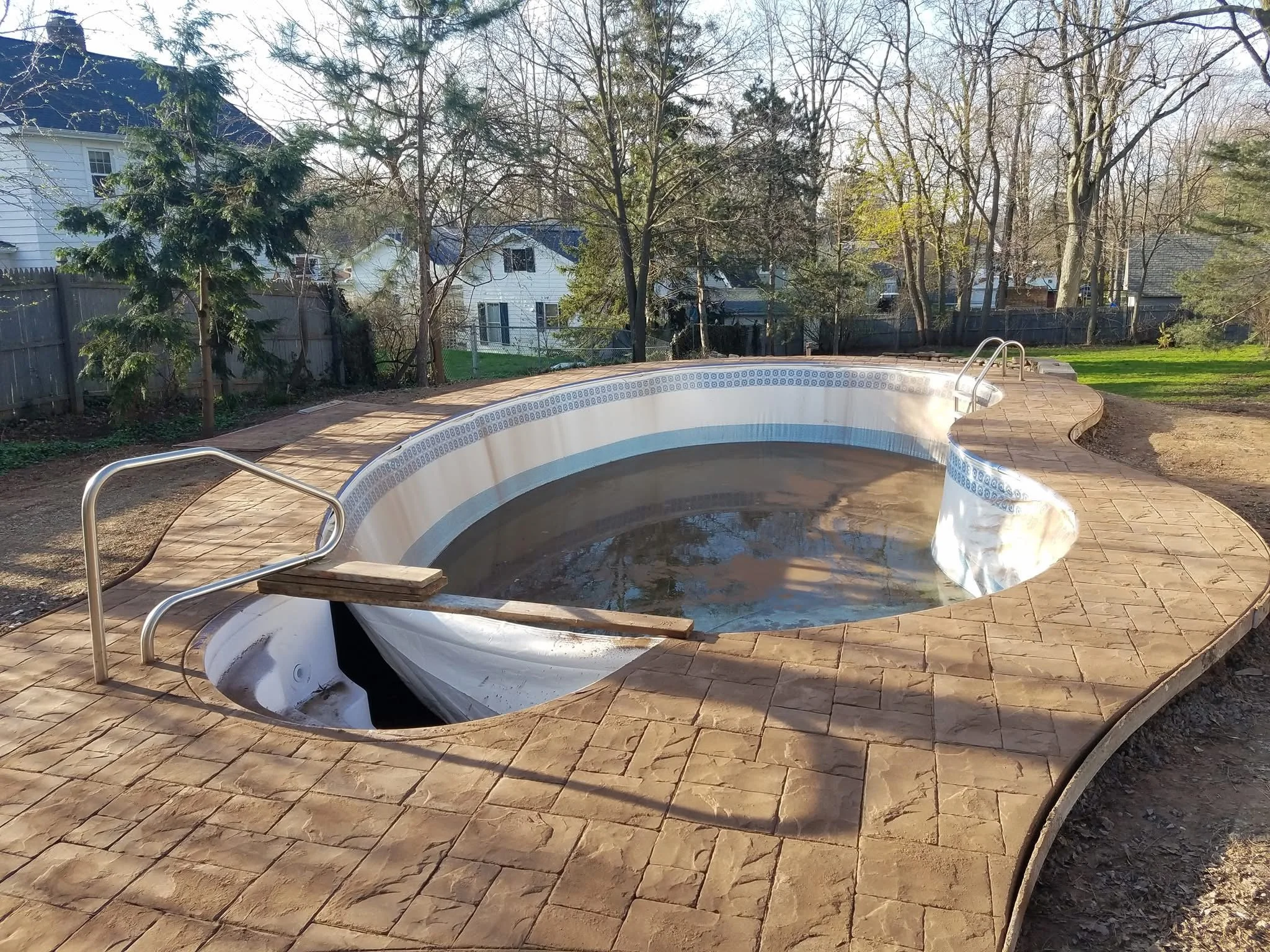 A backyard in the evening with an empty hot tub filled with dirty water, surrounded by a newly laid stamped concrete patio, with trees and houses in the background.