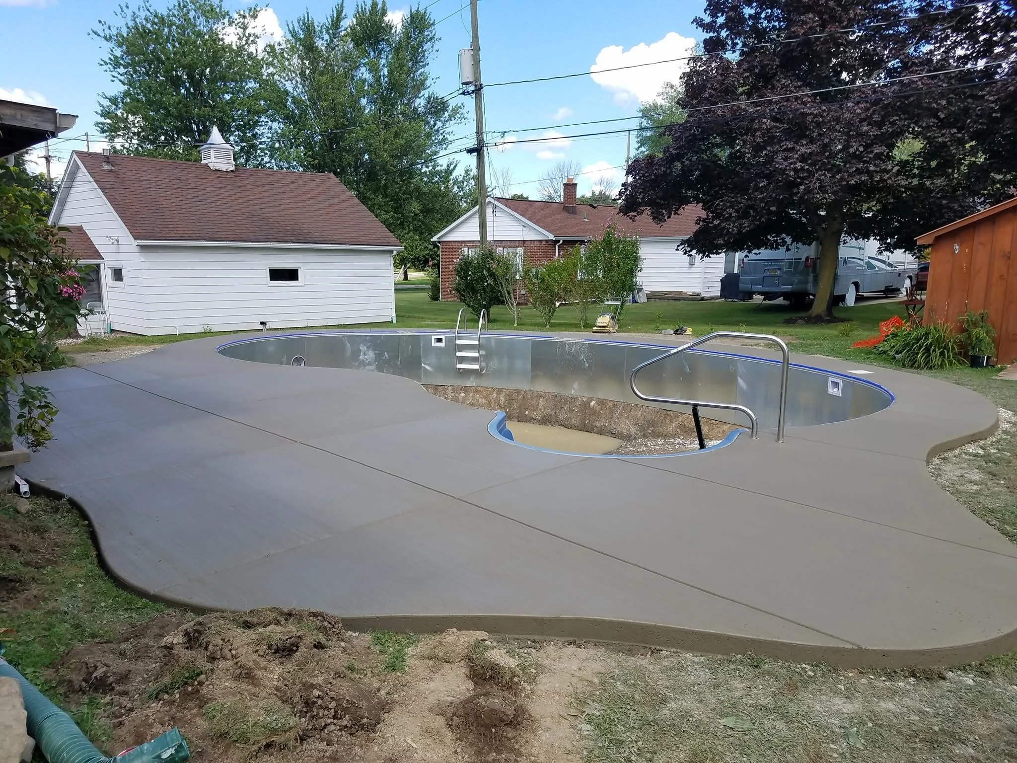 Newly constructed in-ground swimming pool with metal sides and steps, surrounded by a smooth concrete deck in a backyard with trees, small buildings, and a RV in the background.