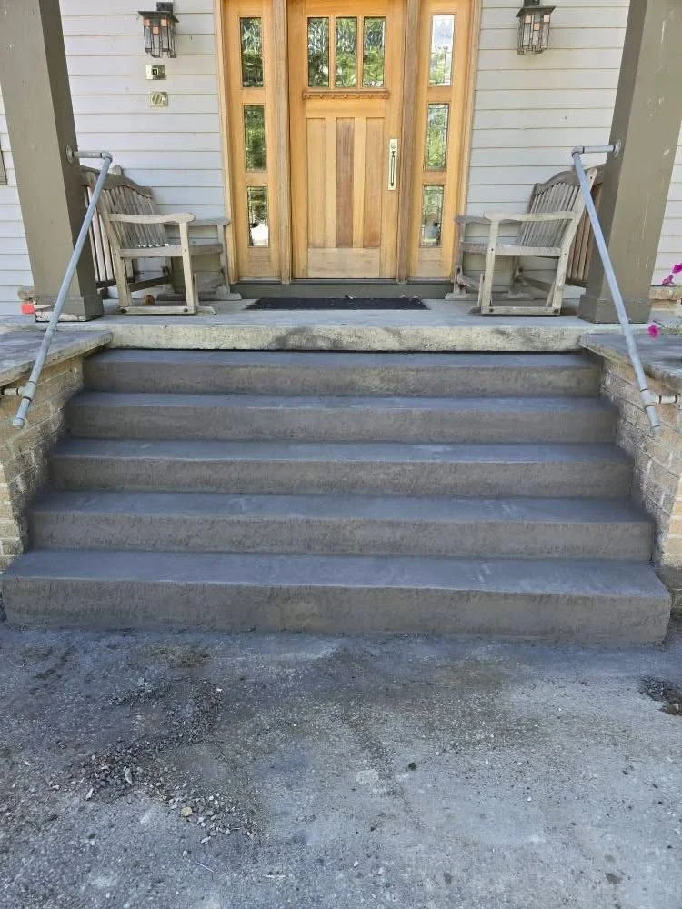 Concrete stairs leading up to a front door with a wooden door and sidelights, flanked by two benches and two outdoor wall lights.