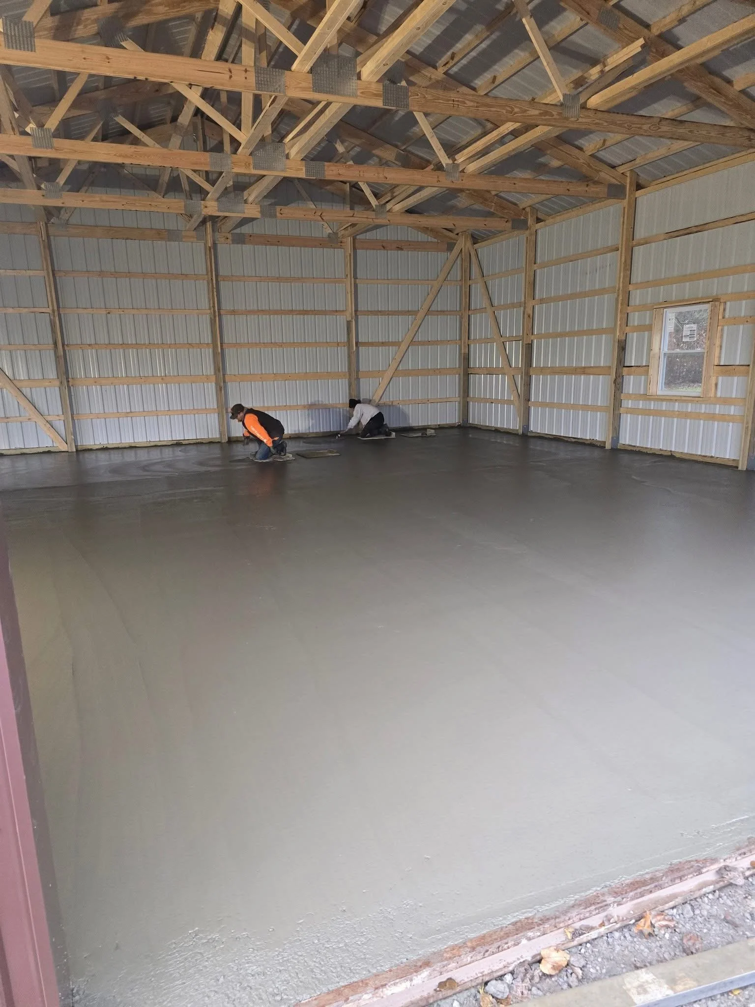 Two workers are kneeling on the floor of a large, open barn, smoothing freshly poured concrete. The barn has unfinished wooden framing and metal siding, with a window on the right wall.
