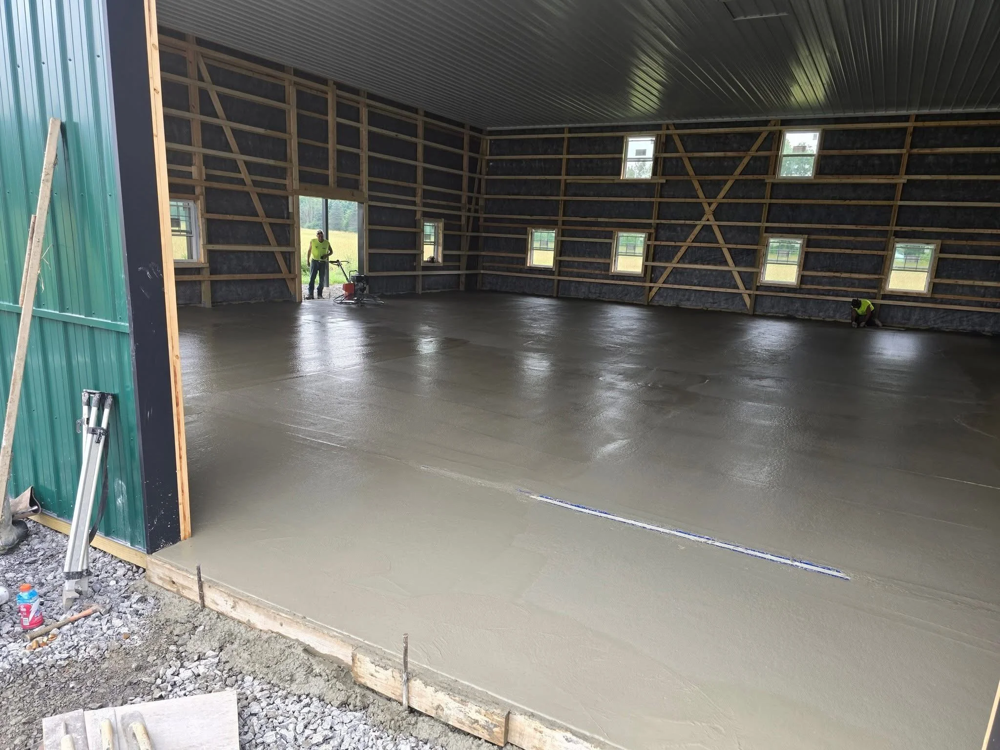 Interior of a building under construction with workers smoothing wet concrete on the floor, wooden framing visible on the walls, multiple small windows, and a partially open side showing outdoor scenery.