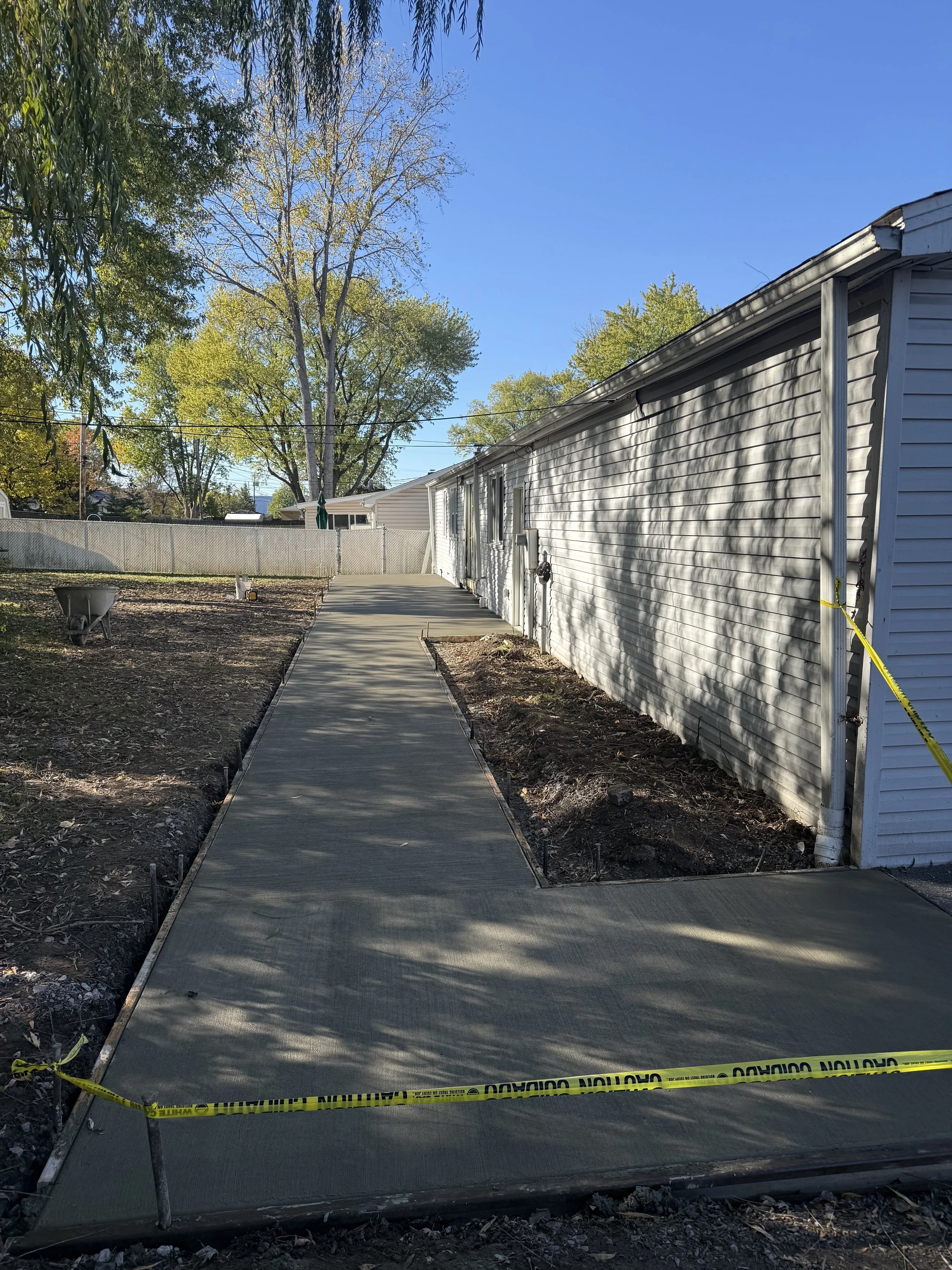 Newly poured concrete sidewalk running alongside a white house, with caution tape blocking the section. There is a yard on one side with some bare soil and trees in the background.