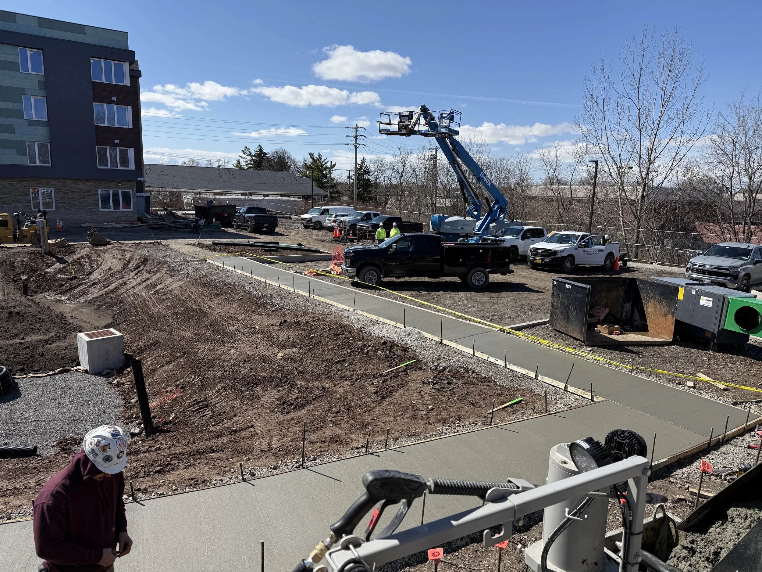 Construction site with workers, trucks, and a lift, paving pathways around a building with a blue sky and trees in the background.