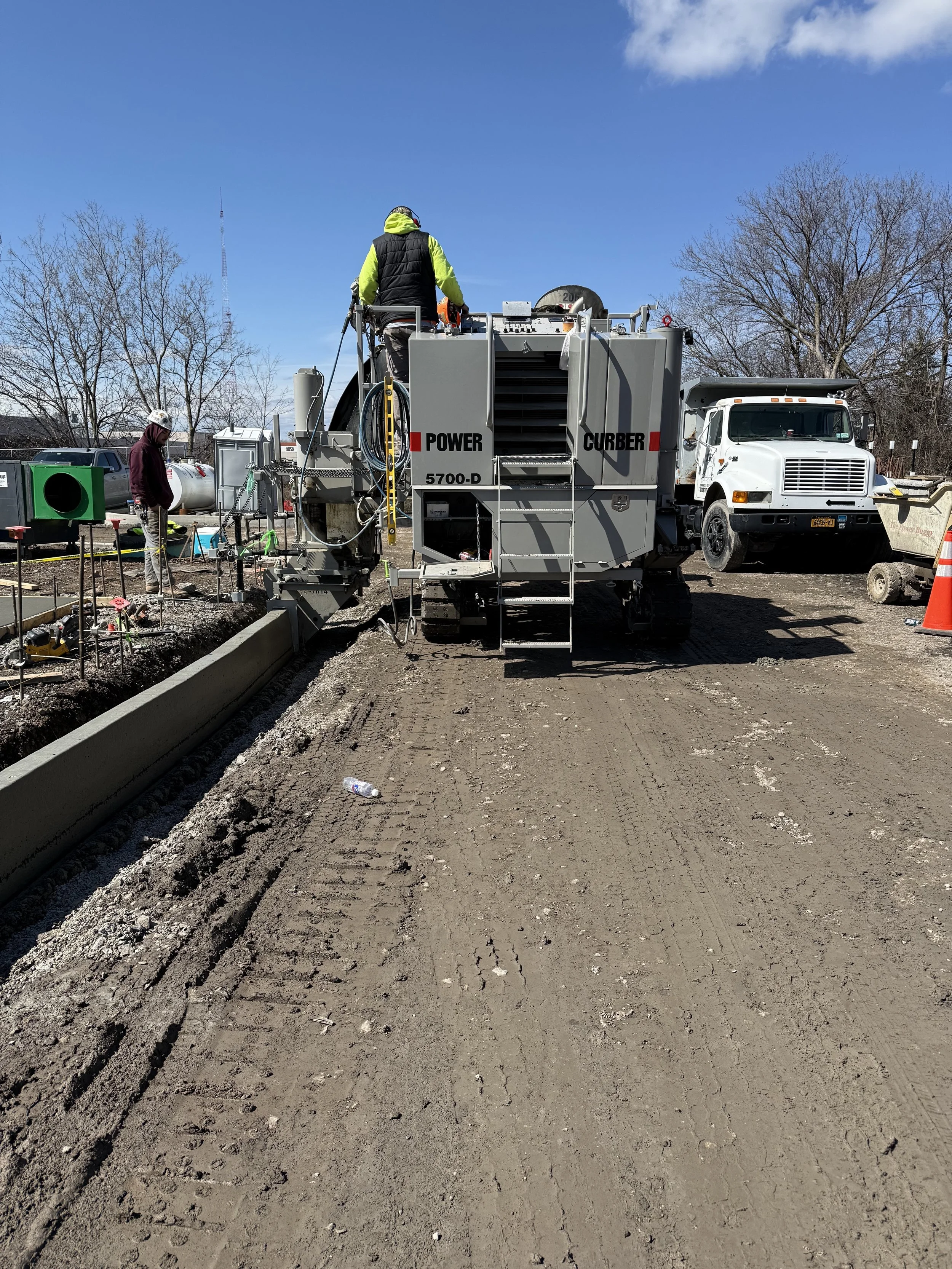 Construction workers operate a large road milling machine on a dirt road during daytime, with trees and vehicles in the background.