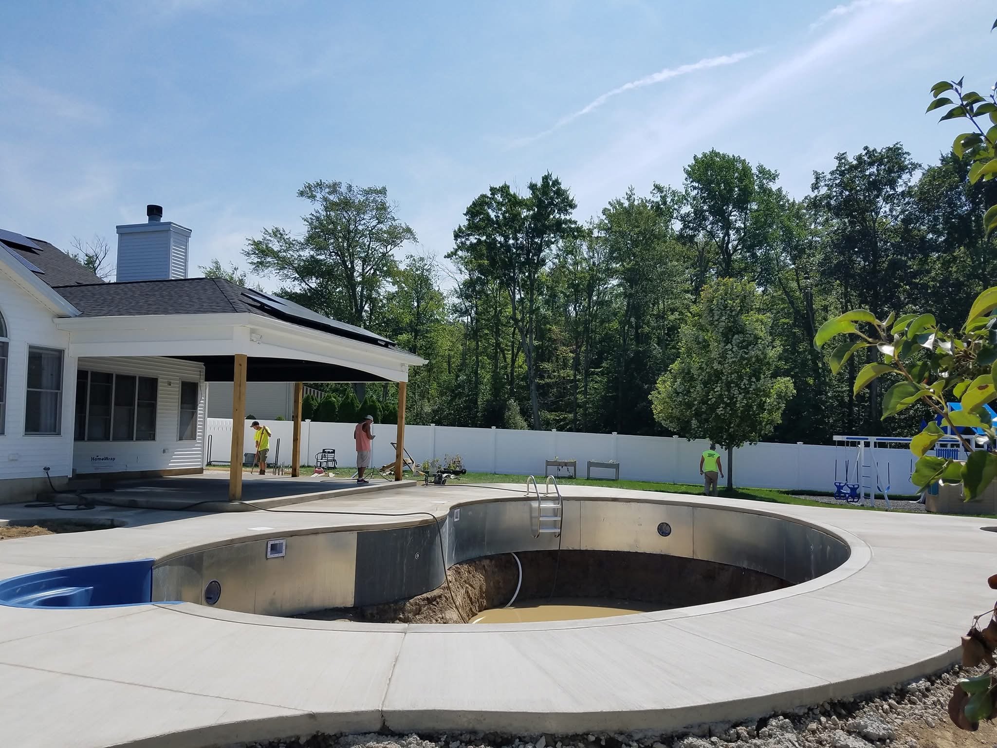 In-ground swimming pool under construction in backyard with workers, surrounded by white fence, trees, and a house with a covered patio.