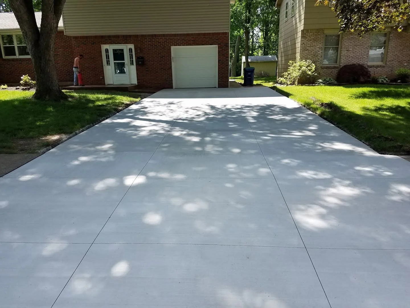 Freshly poured concrete driveway leading to a house with a brick exterior, white garage door, and a person watering plants near the front door, with green lawns and trees on either side.