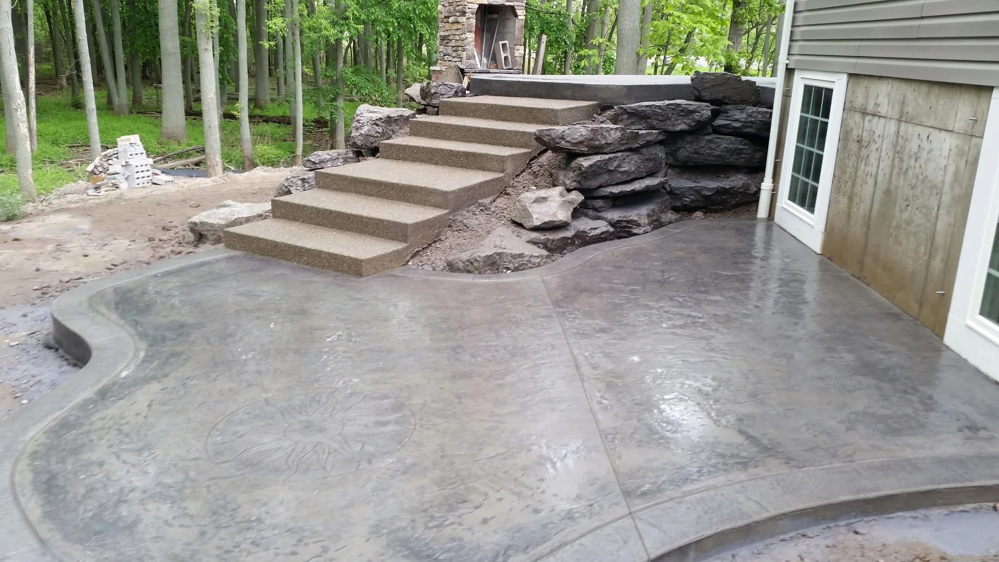 Concrete patio with curved edge, adjacent to a house with sliding glass doors. Stone steps lead up to a wooded area with trees and grass in the background.