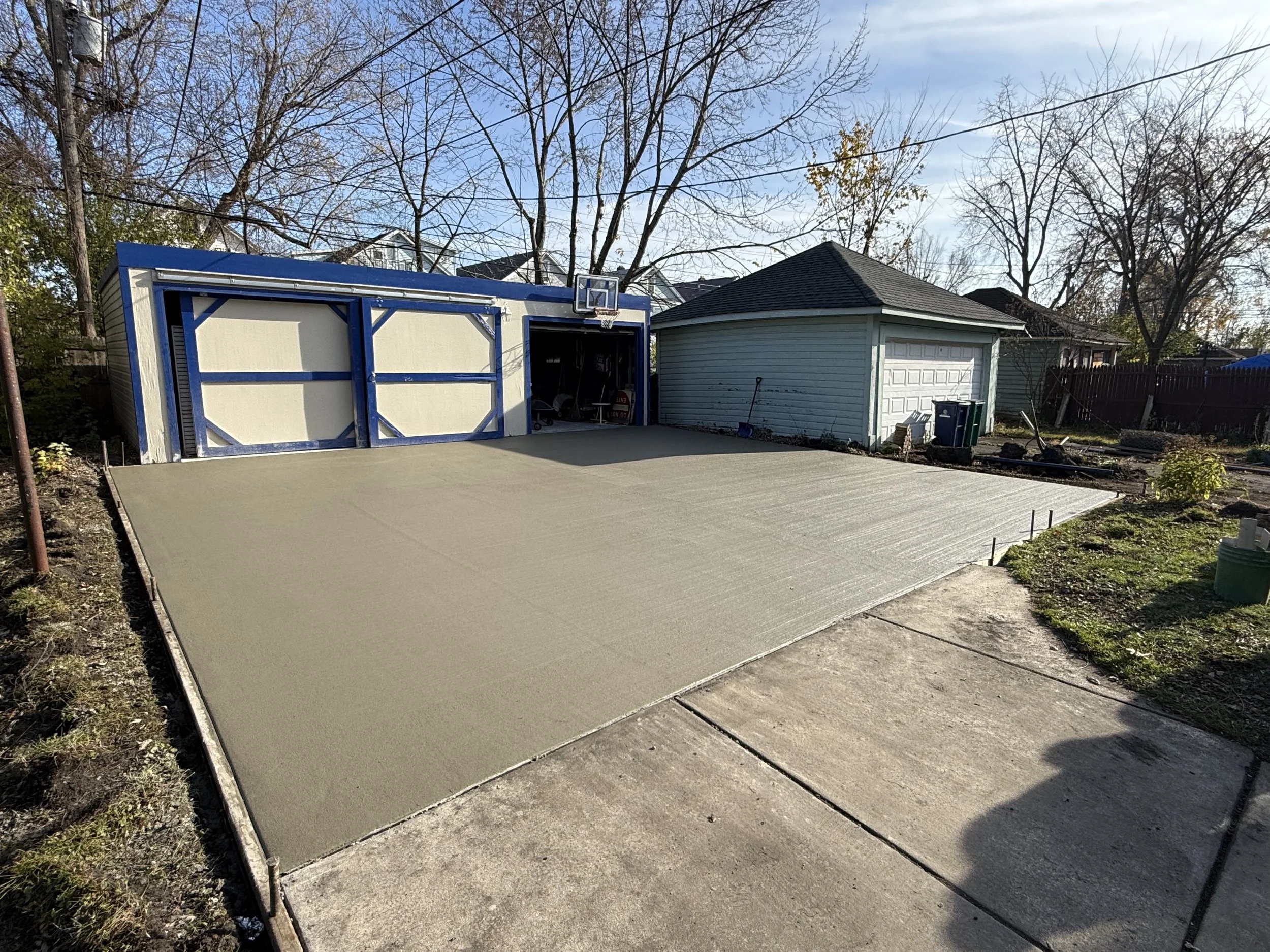Freshly poured concrete driveway in front of a garage during daytime, with trees and a shed visible in the background.