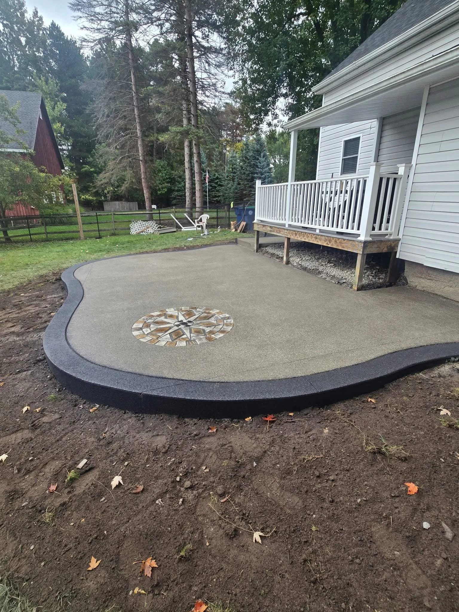 Newly poured concrete patio with a decorative circular inlay in the center, adjacent to a house with a small porch, surrounded by a backyard with trees and a fence.