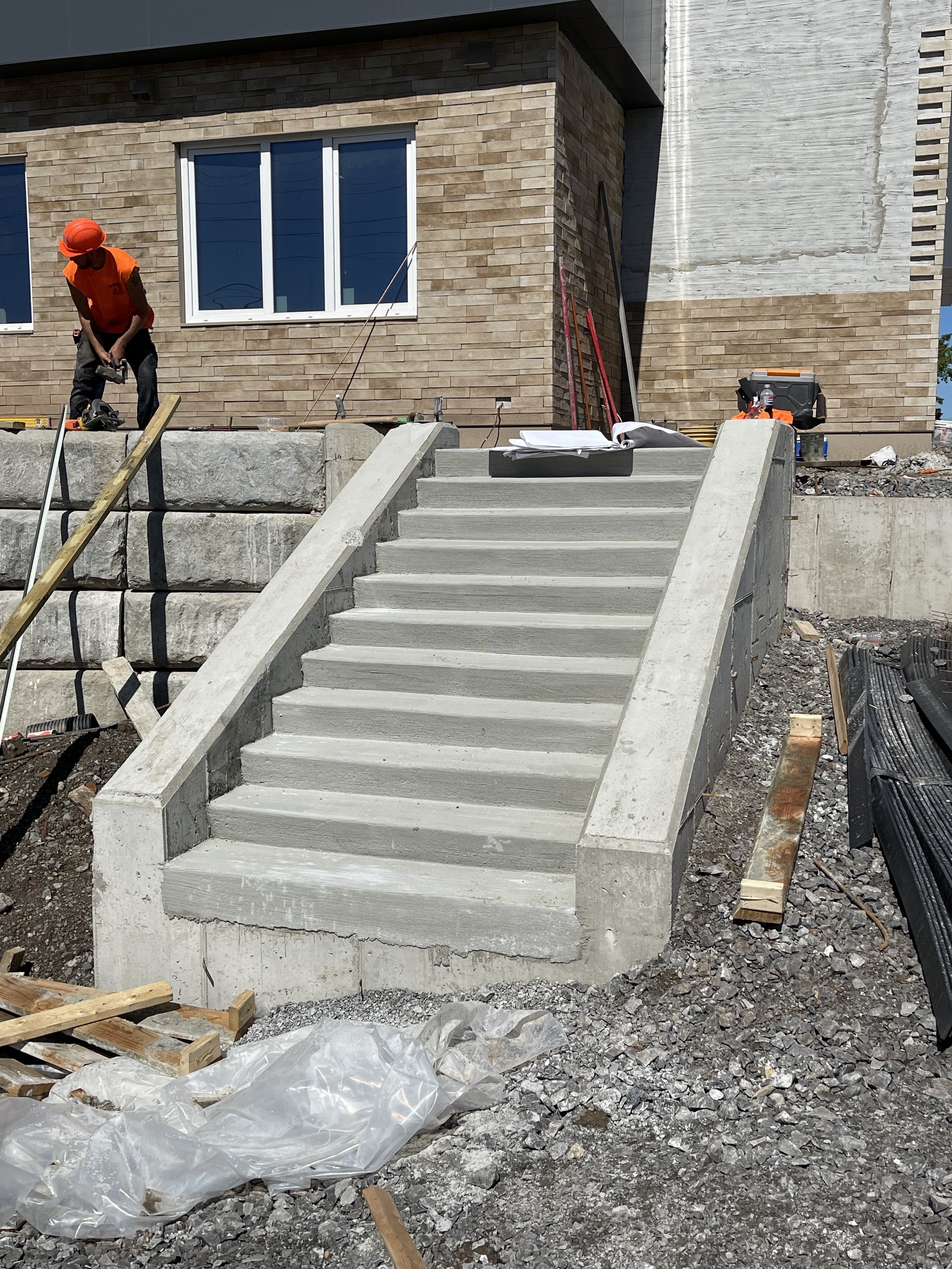 Construction workers building concrete stairs outside a modern house with brick siding, construction tools, and materials around.