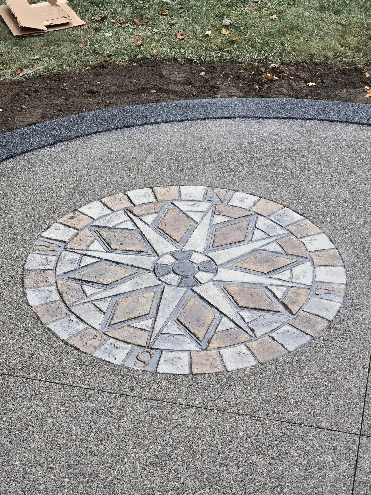 Concrete and tile compass rose on sidewalk with grass and fallen leaves in background.