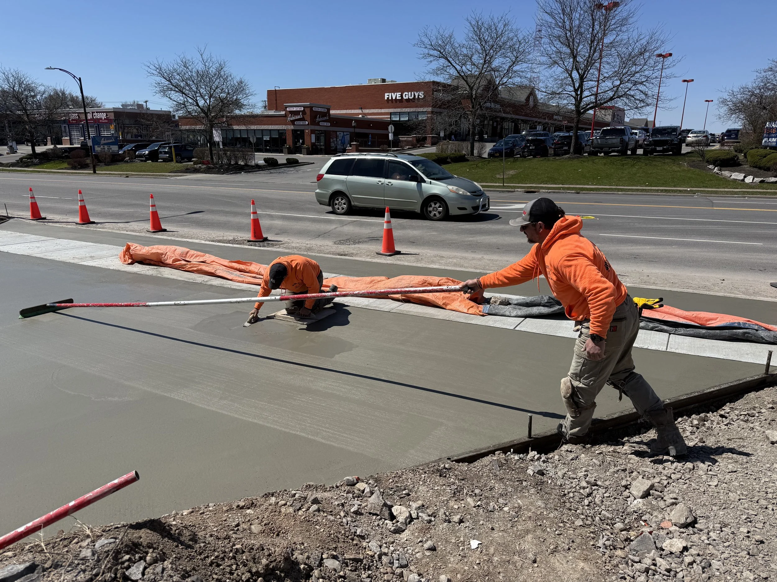 Construction workers in orange hoodies pouring and leveling concrete on a sidewalk in an urban area with traffic cones, a silver minivan, and restaurants in the background.