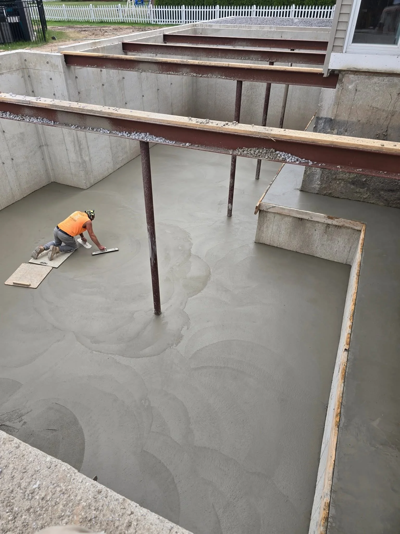 Construction worker leveling freshly poured concrete in a building foundation with unoccupied concrete walls and steel beams overhead.