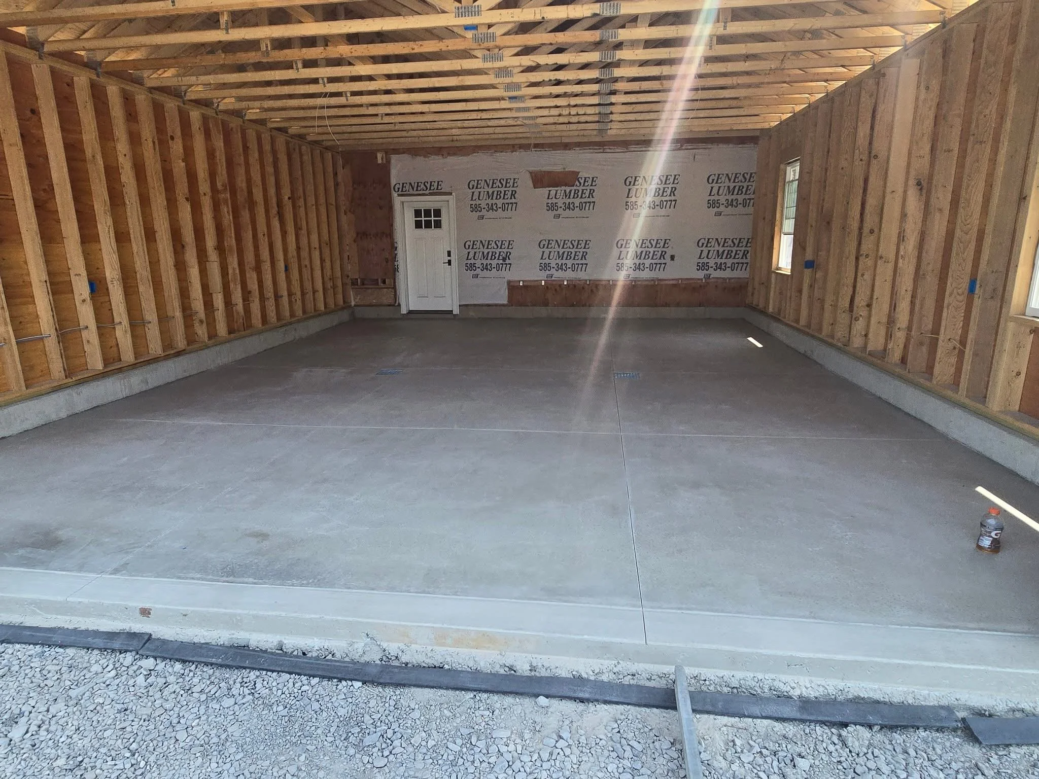 Interior view of a garage under construction with exposed wooden framing, concrete floor, and a door with a window on the back wall. The walls are partially covered with construction signs for 'Genesee Lumber,' and multiple windows are on the side wa