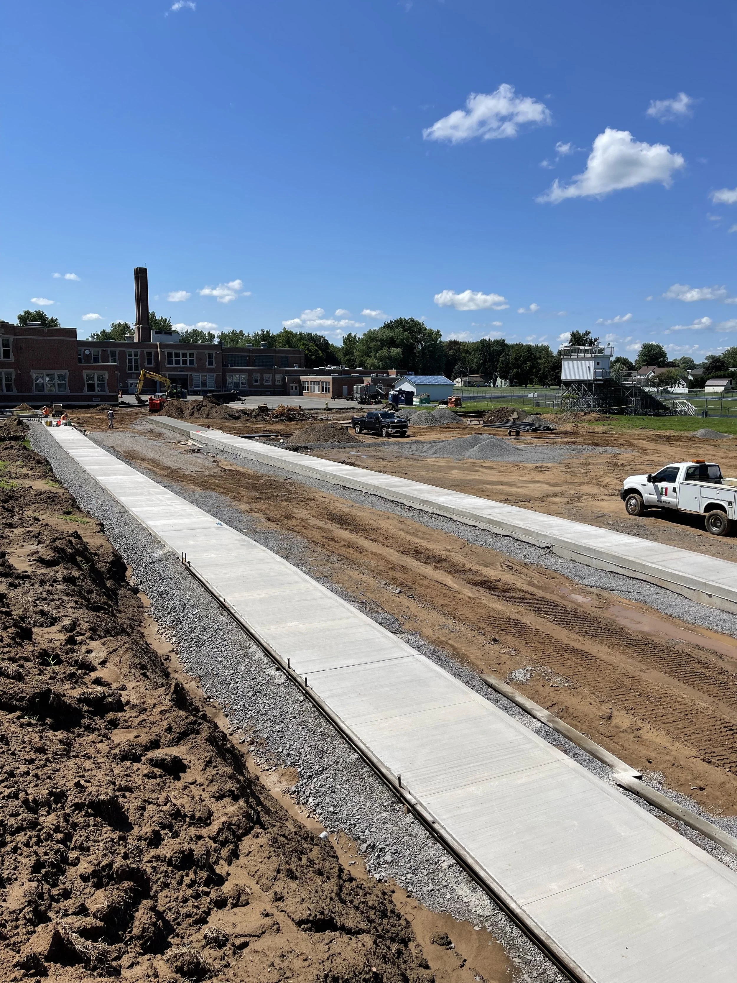 Construction site with concrete sidewalk being built, trucks and construction vehicles on muddy ground under a blue sky with clouds.