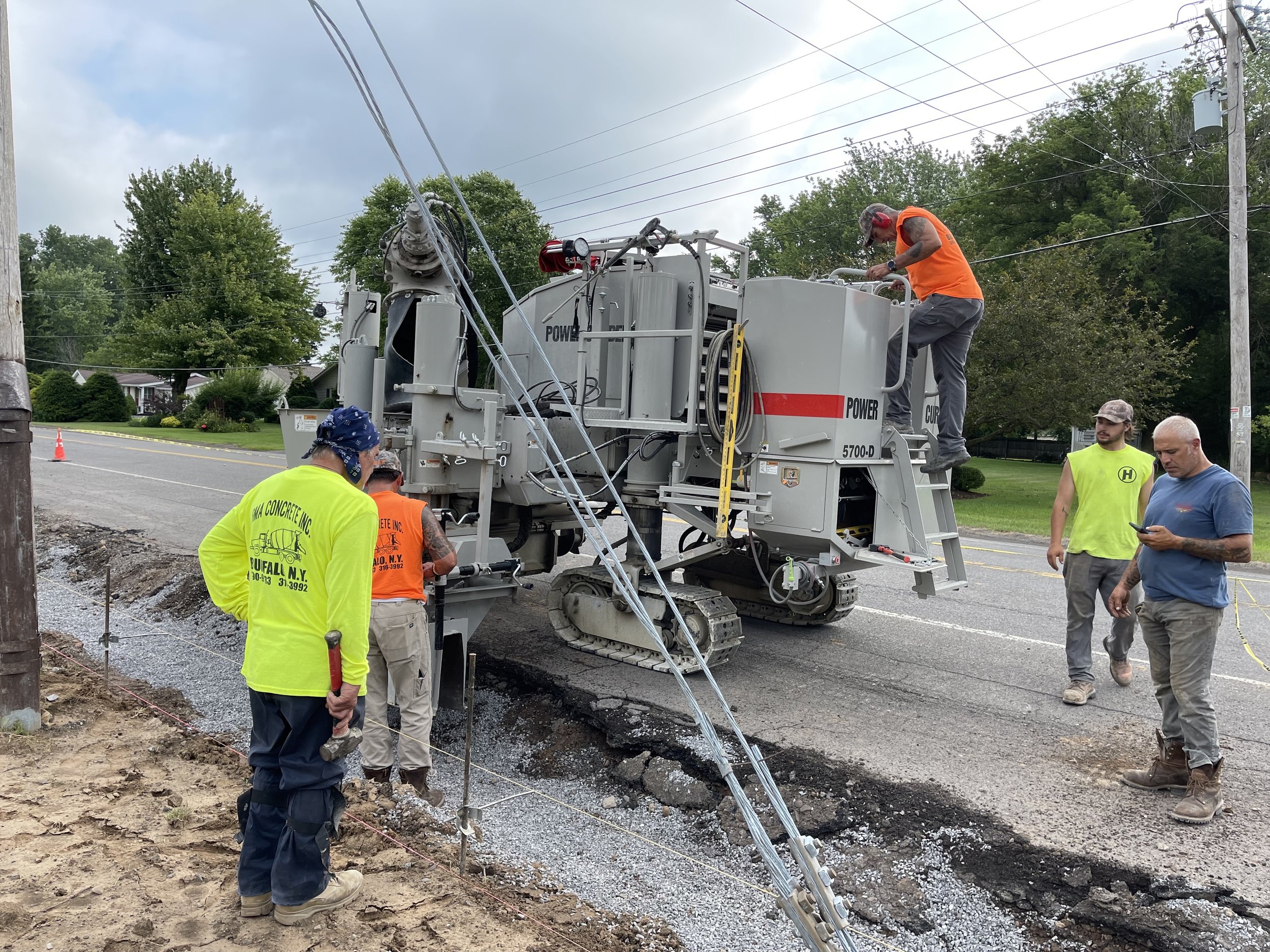 Construction workers operate machinery and inspect a road repair site on a street, with utility poles and trees in the background.