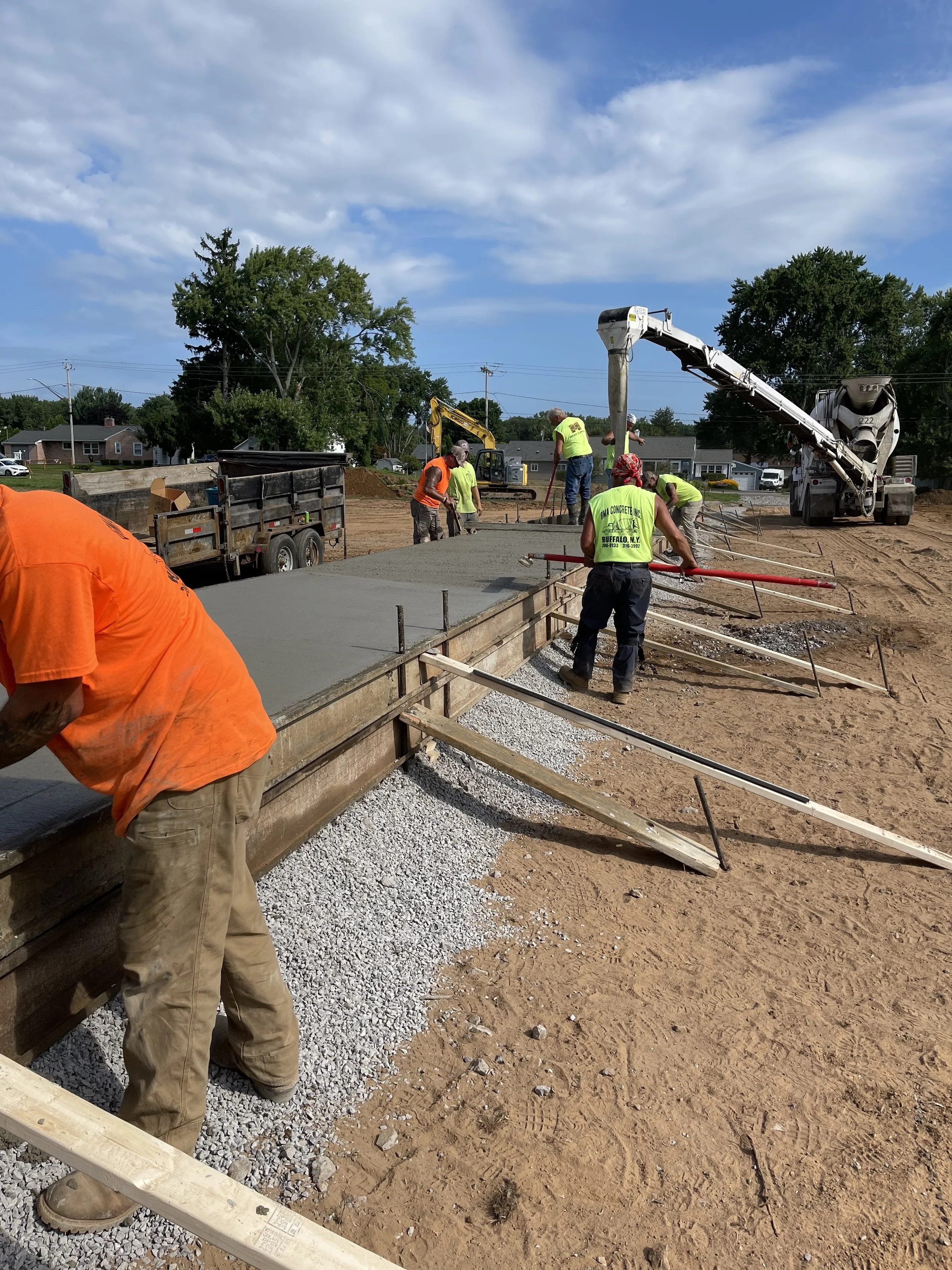Construction workers pouring and spreading concrete on a building foundation with construction equipment and support structures, surrounded by a dirt area and a partly cloudy sky.