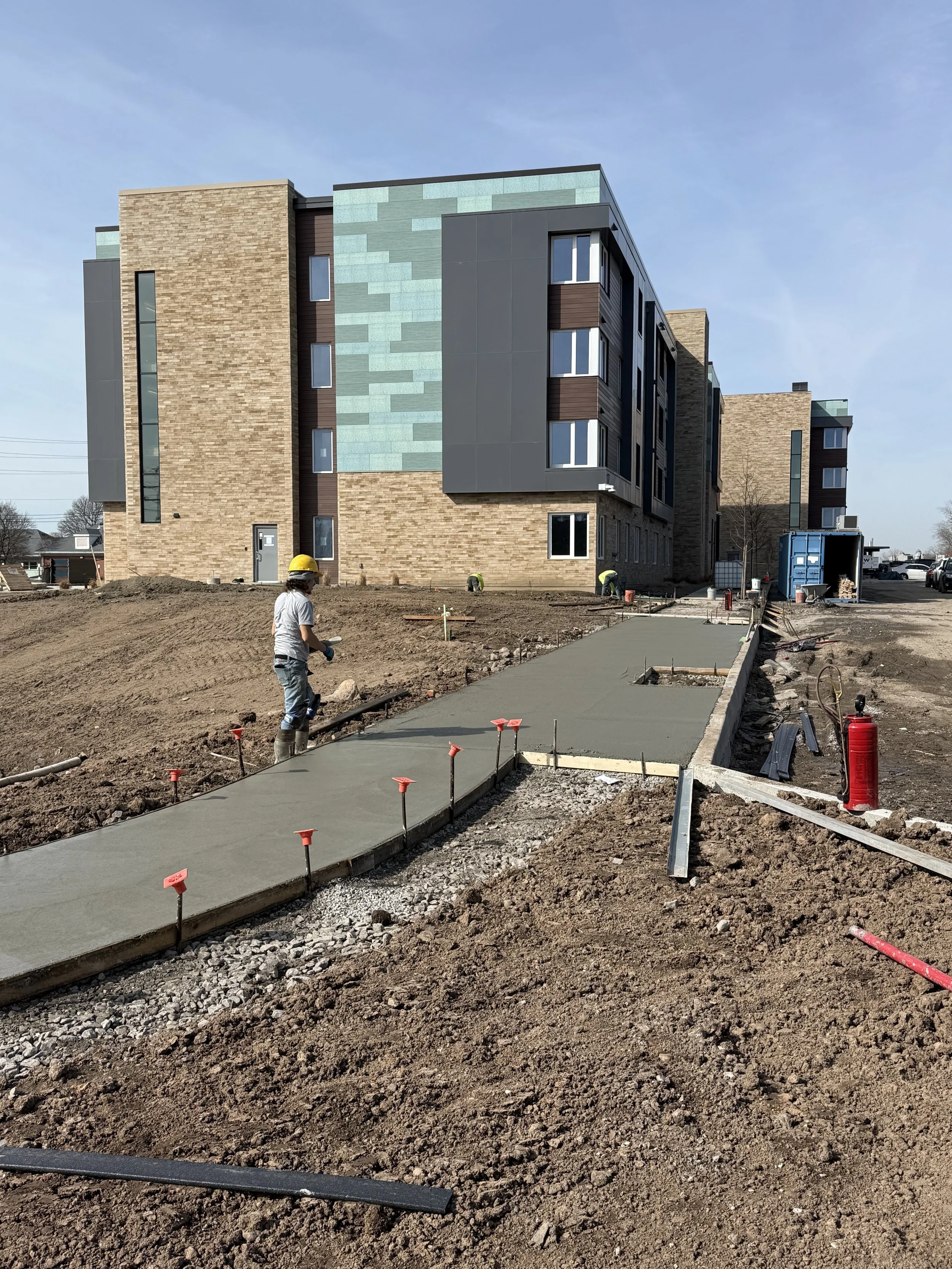 Construction workers are pouring a concrete sidewalk in front of a multi-story residential building. The building has brick, blue, and dark gray exterior panels. Construction supplies and equipment are visible around the site.
