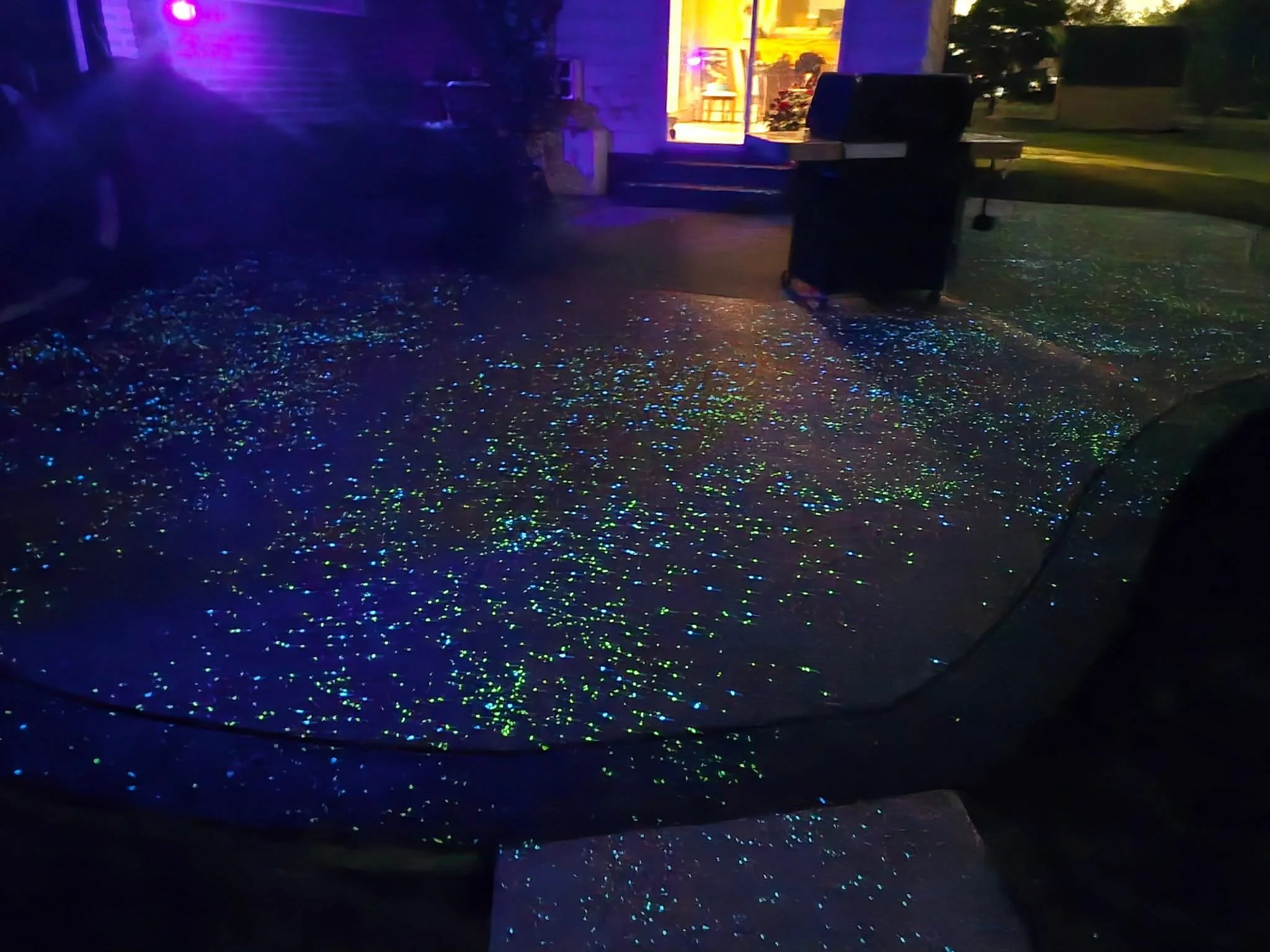 Nighttime outdoor scene showing a patio with glowing, multicolored glitter scattered on the ground, an open door revealing a brightly lit interior, and a barbecue grill nearby.