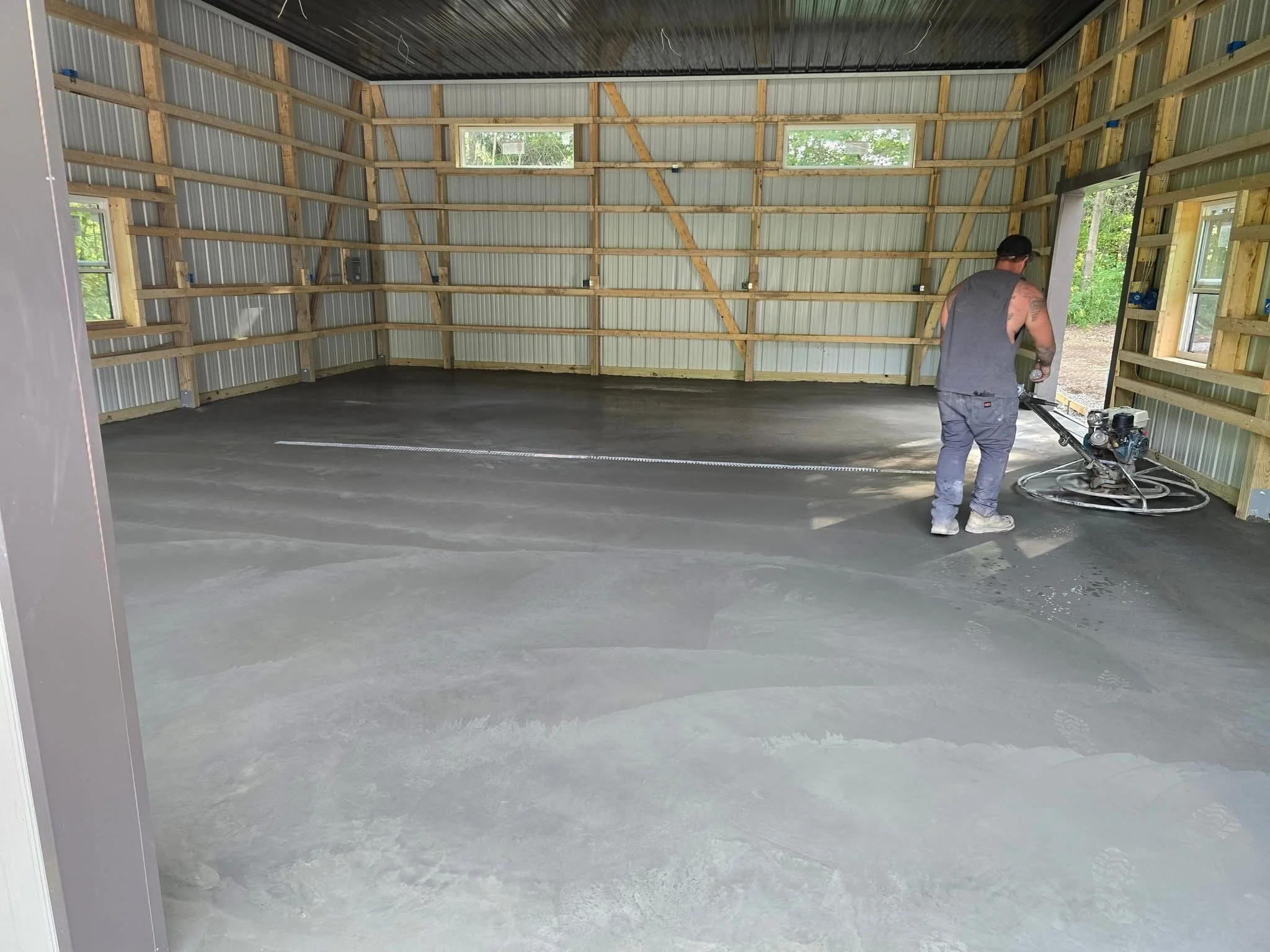 A man working with a concrete power trowel machine inside a partially built garage with metal walls and wooden framing, and multiple windows.