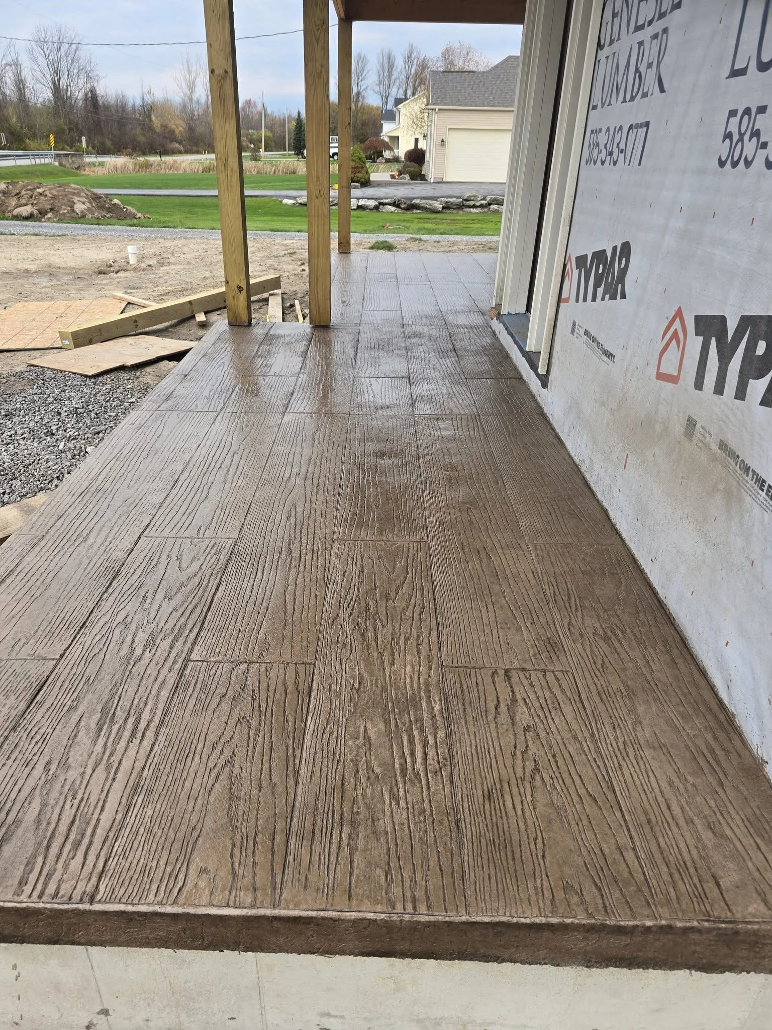 A newly installed wooden deck outside a house under construction, with decorative textured wood grain pattern on the floor. The house has an unfinished wall with building wrap labeled 'TYPAR' and some wooden support beams.