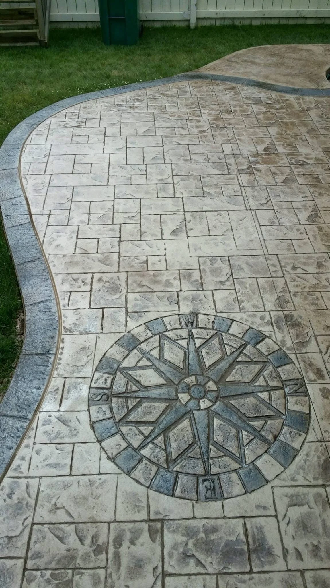Concrete backyard patio with a compass rose design etched into the surface, surrounded by a curved stone border and a small grassy area with a fence in the background.