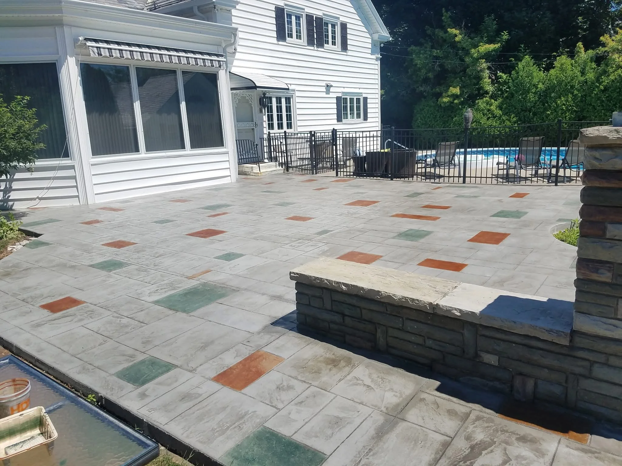 A spacious backyard with a newly paved stone patio, surrounded by a black metal fence, a white house with black shutters in the background, and a swimming pool with lounge chairs beyond the fence.
