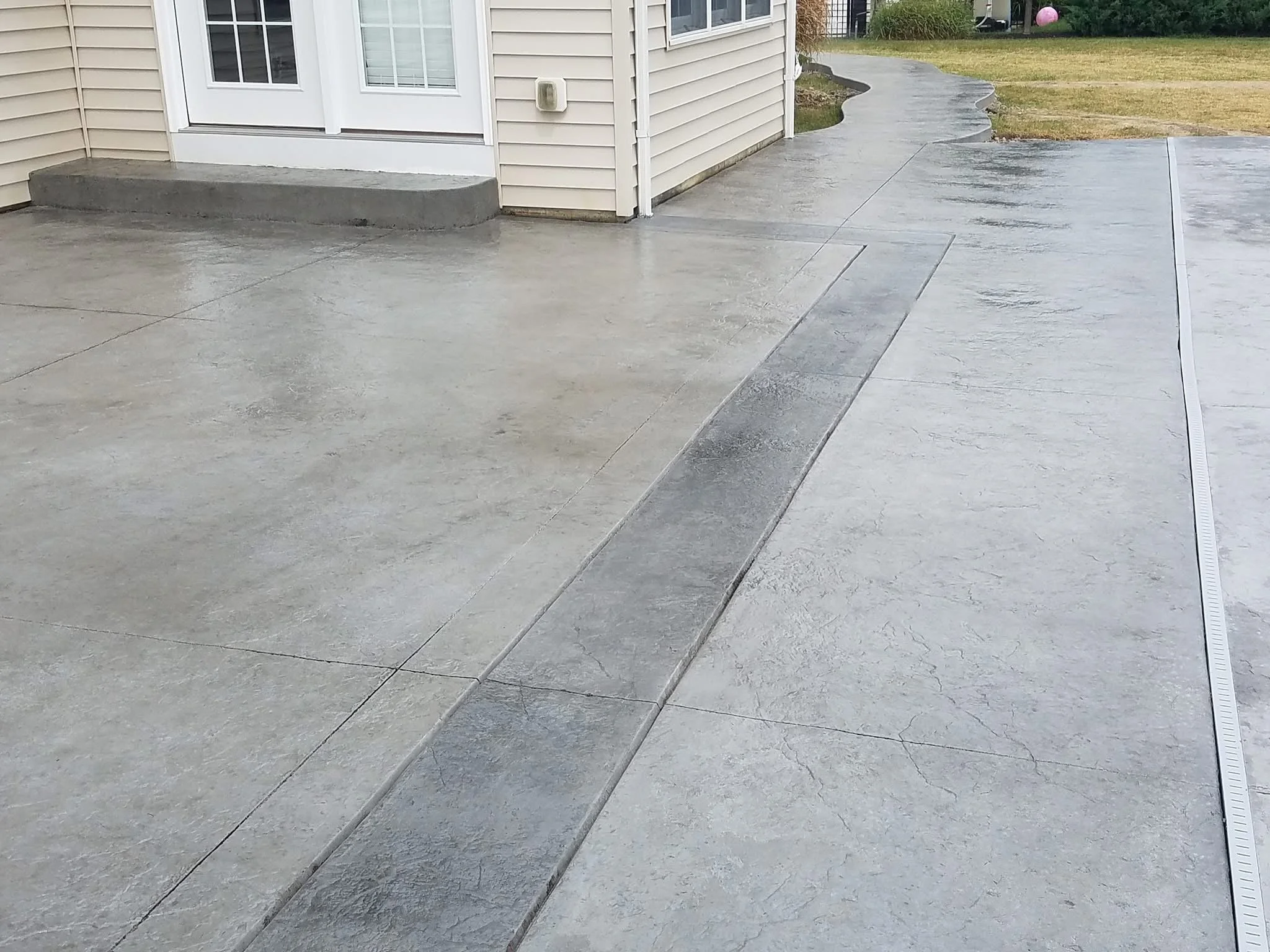 Wet concrete sidewalk with a drainage grate on the right side and a house with siding, a small set of steps, and a window in the background.