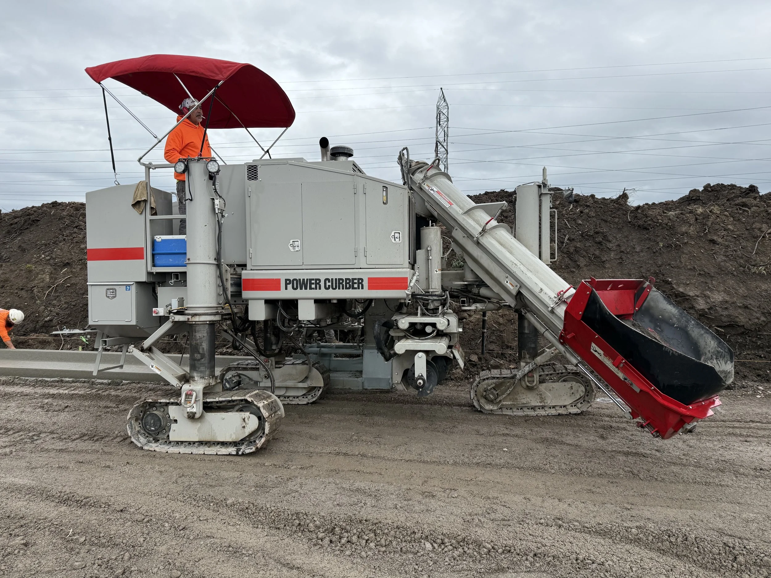 Construction worker operating a large power curber machine with tracks and a conveyor belt on a dirt road, overcast sky.