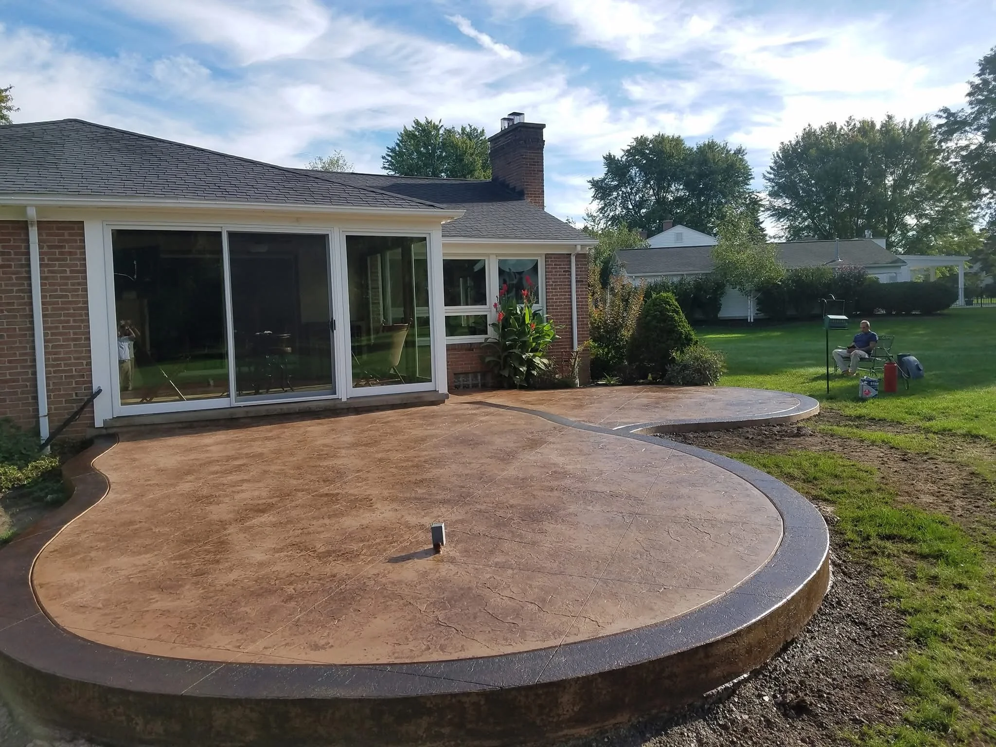 Backyard with newly poured concrete patio, brick house with sliding glass door, house with chimney, man sitting on a bench, gardening tools and grass.