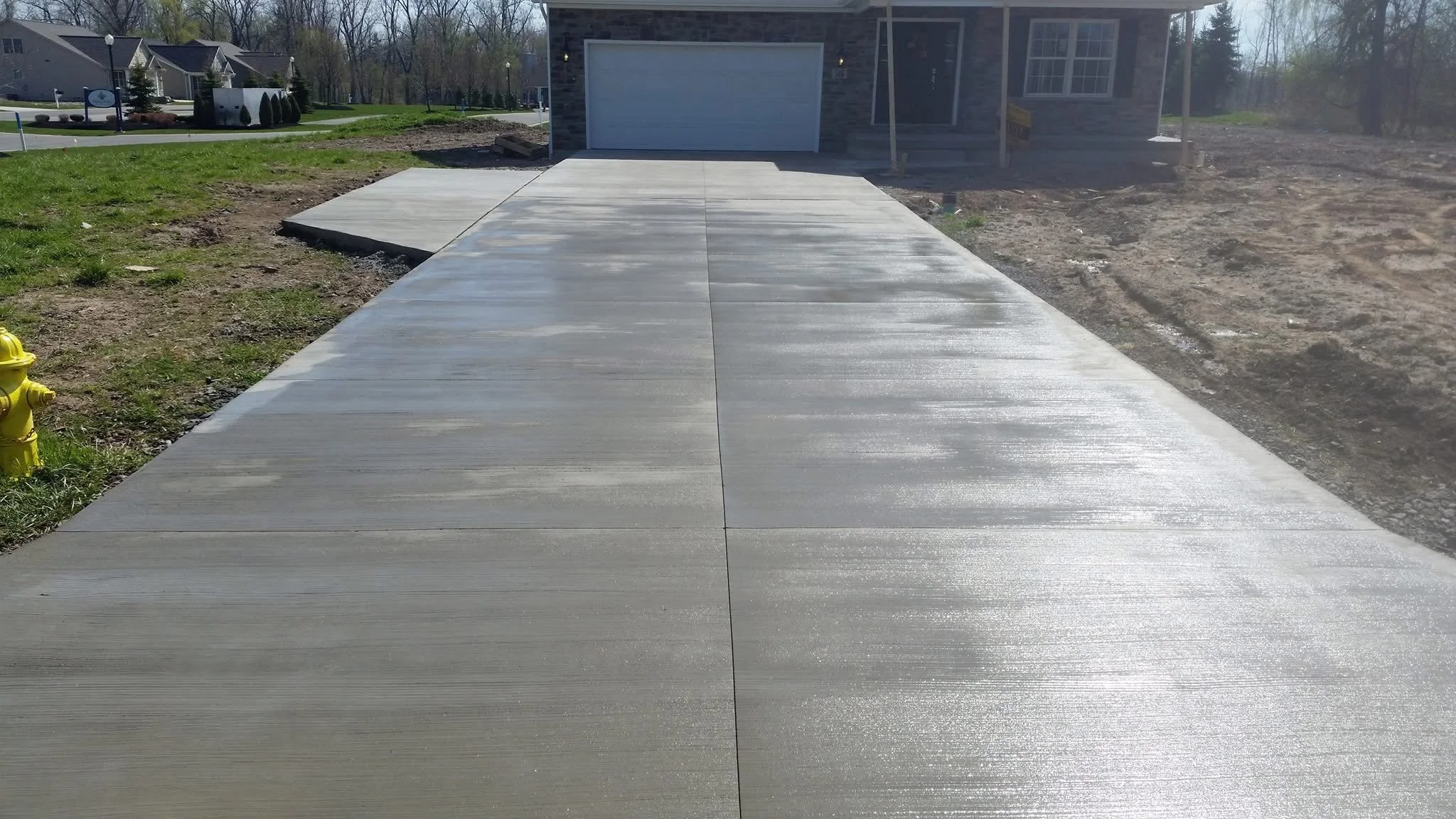 Newly poured concrete sidewalk leading to a house with a garage, surrounded by a grassy area on the left and dirt on the right, with neighboring houses visible in the background.