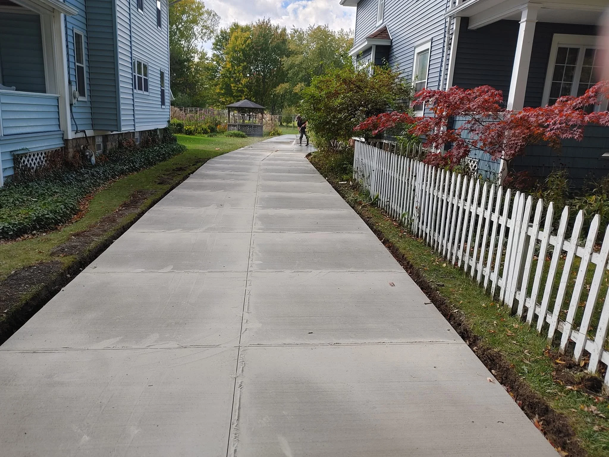 Concrete sidewalk in a residential neighborhood with houses on both sides, a white picket fence, plants, and trees with green and red leaves, and a gazebo in the background.