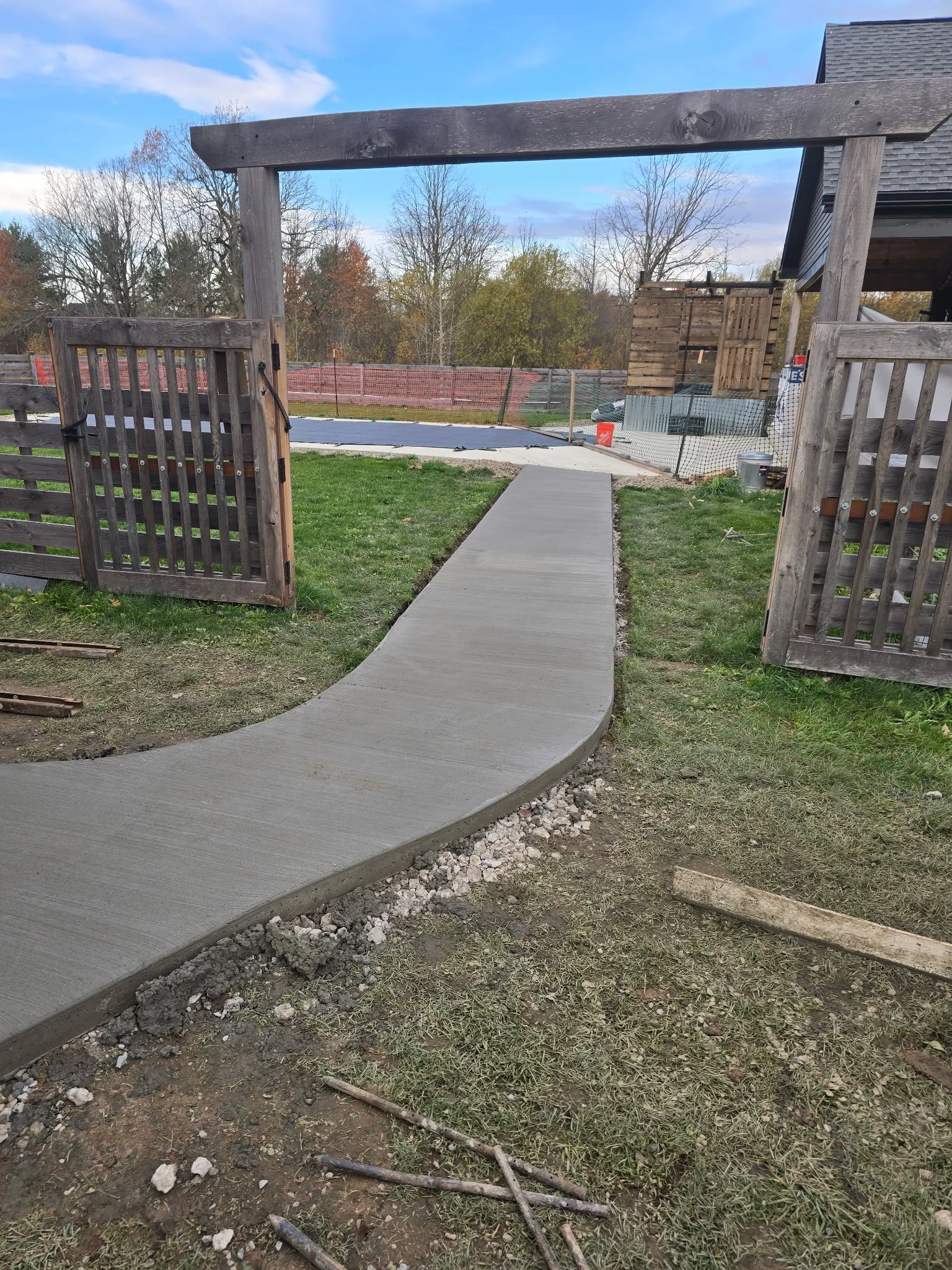 Newly poured concrete sidewalk leading through a wooden gate toward a fenced backyard with a trampoline and a shed, with autumn trees in the background.
